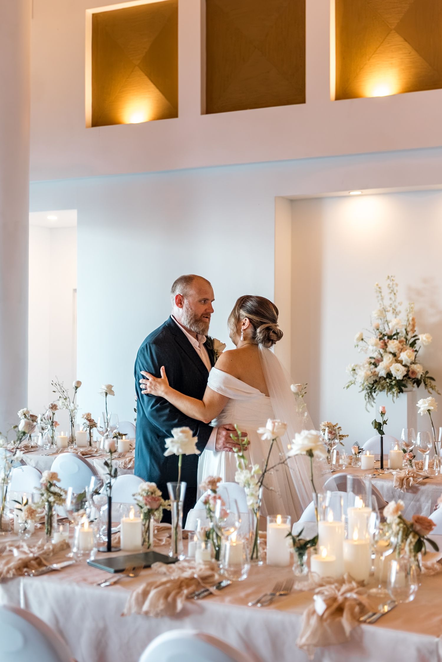 Bride and groom share first dance surrounded by elegant candlelit reception tables with white floral arrangements.