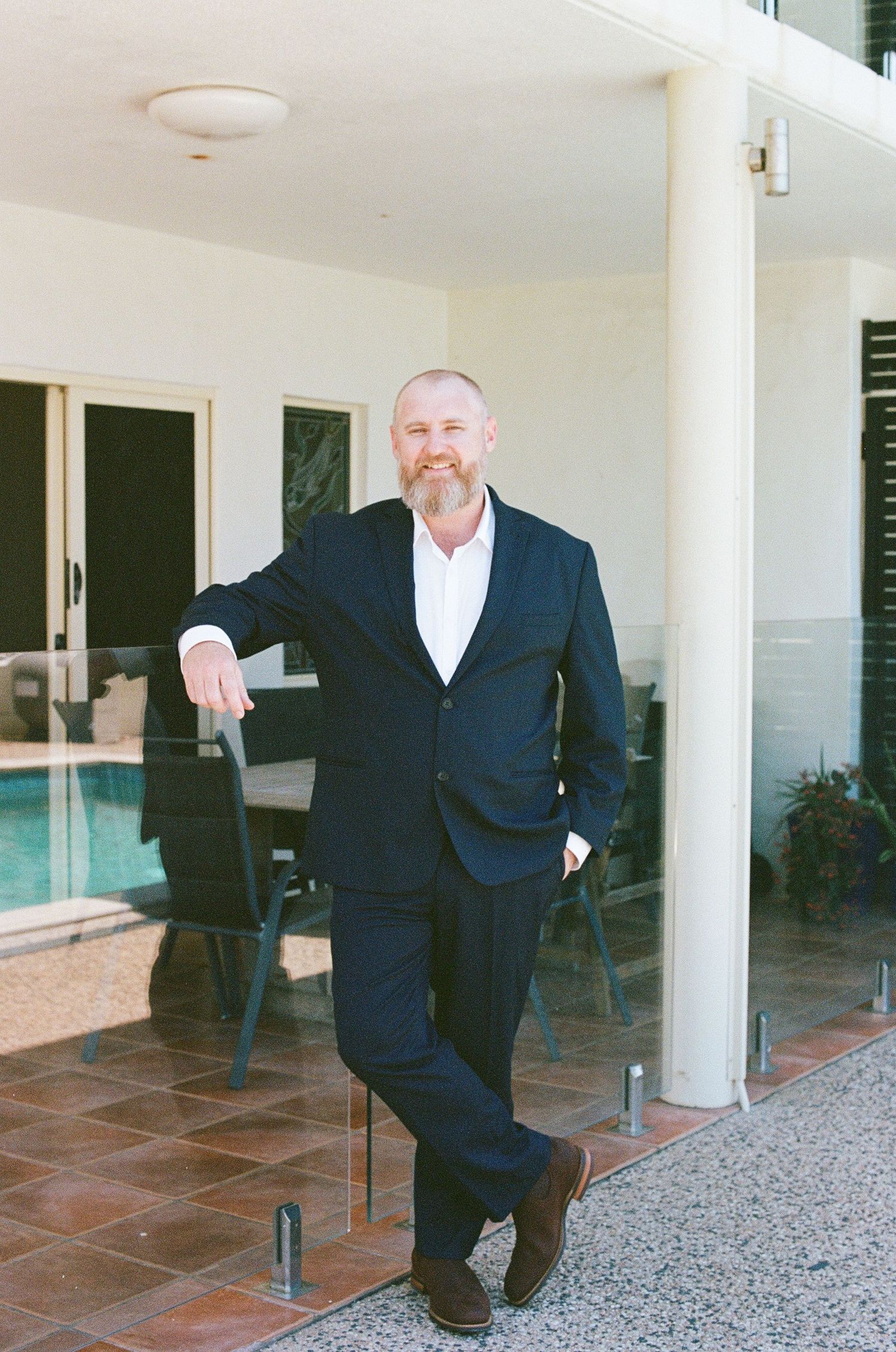 Smiling bearded groom in navy suit leaning against white pillar beside outdoor pool area.