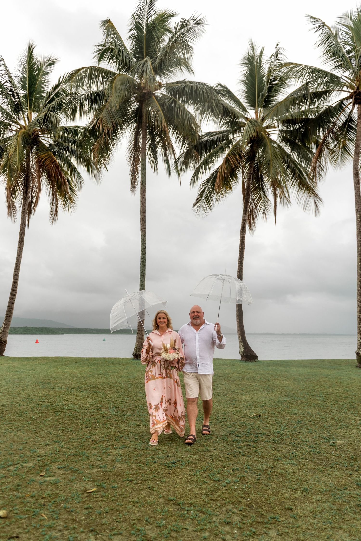 Smiling couple walking toward camera under clear umbrellas on a green lawn lined with tall palm trees.
