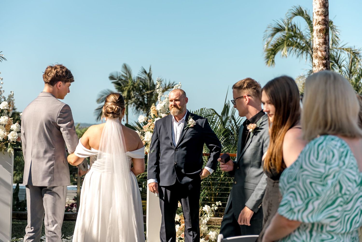 Wedding officiant stands with bride and groom at outdoor tropical ceremony with floral arrangements nearby.