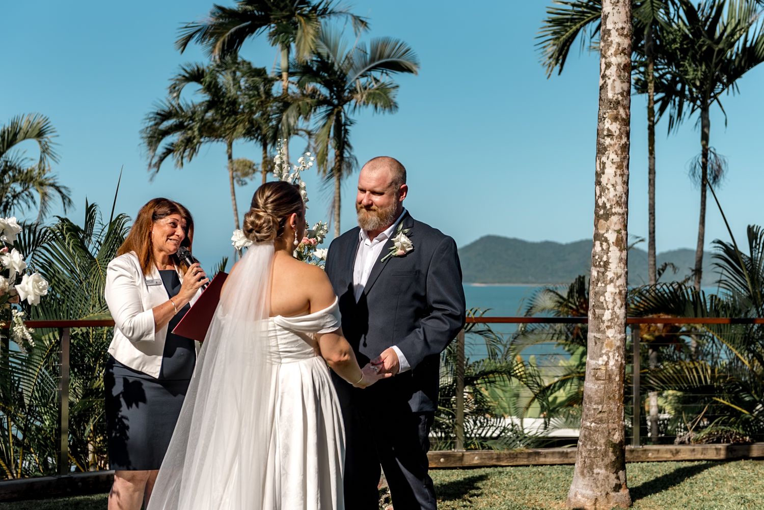 Wedding officiant reads vows as bride and groom hold hands during outdoor tropical ceremony with ocean view.