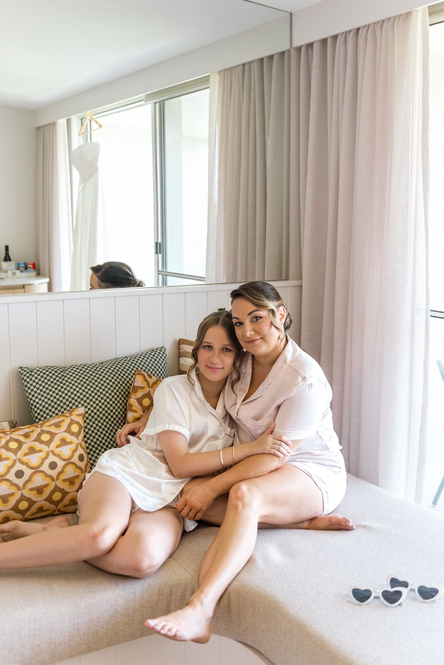 Bride and bridesmaid posing together smiling on a couch in matching silk pajamas before the wedding.