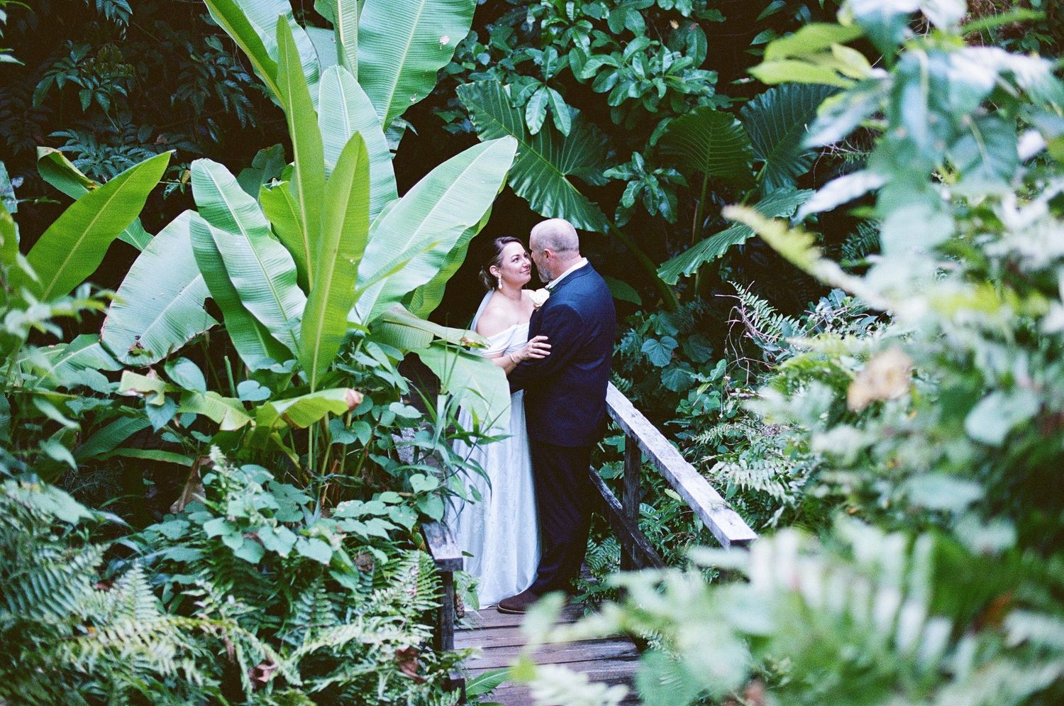 Bride and groom share a romantic embrace on a wooden bridge surrounded by lush tropical greenery.