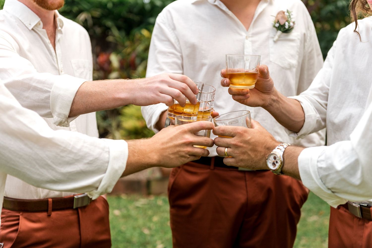 Groomsmen in white shirts and rust-colored pants toasting with whiskey glasses at a wedding celebration.
