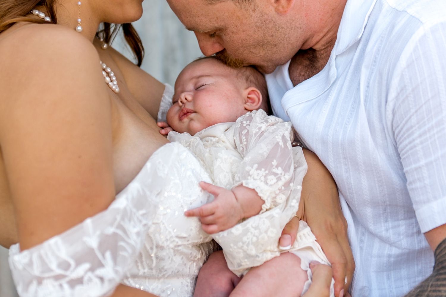 Close-up of bride and groom both kissing their sleeping newborn baby dressed in white lace.