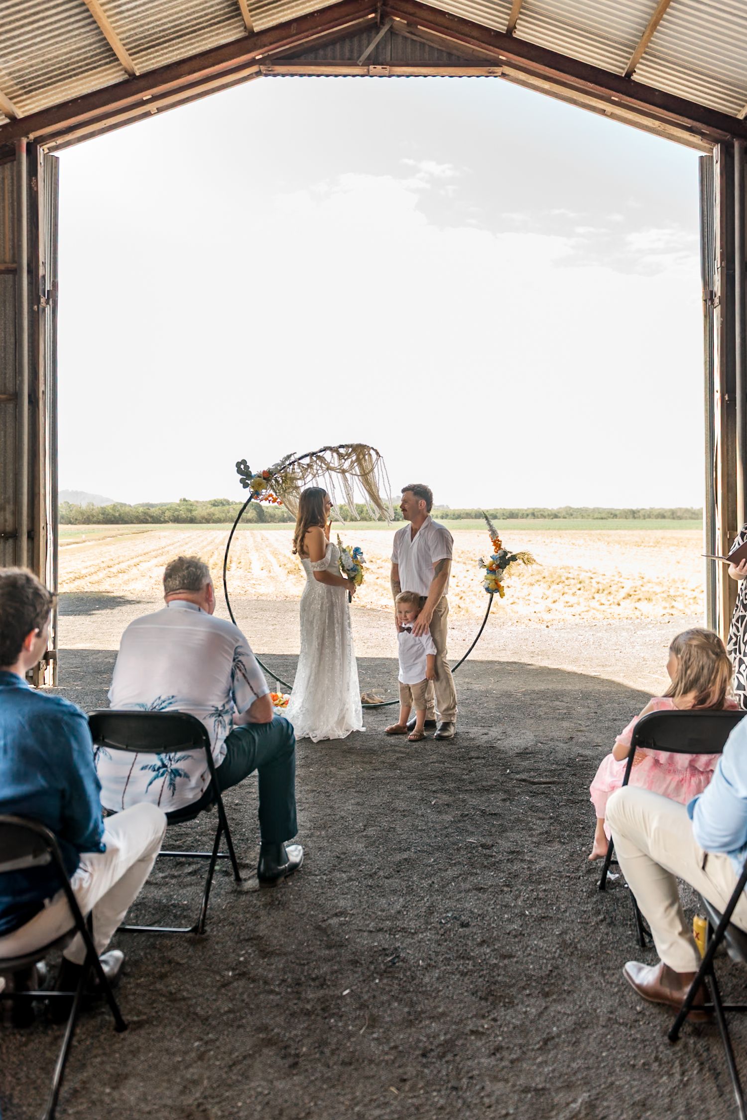 Bride and groom stand with child at circular arch altar inside open barn at sunset.