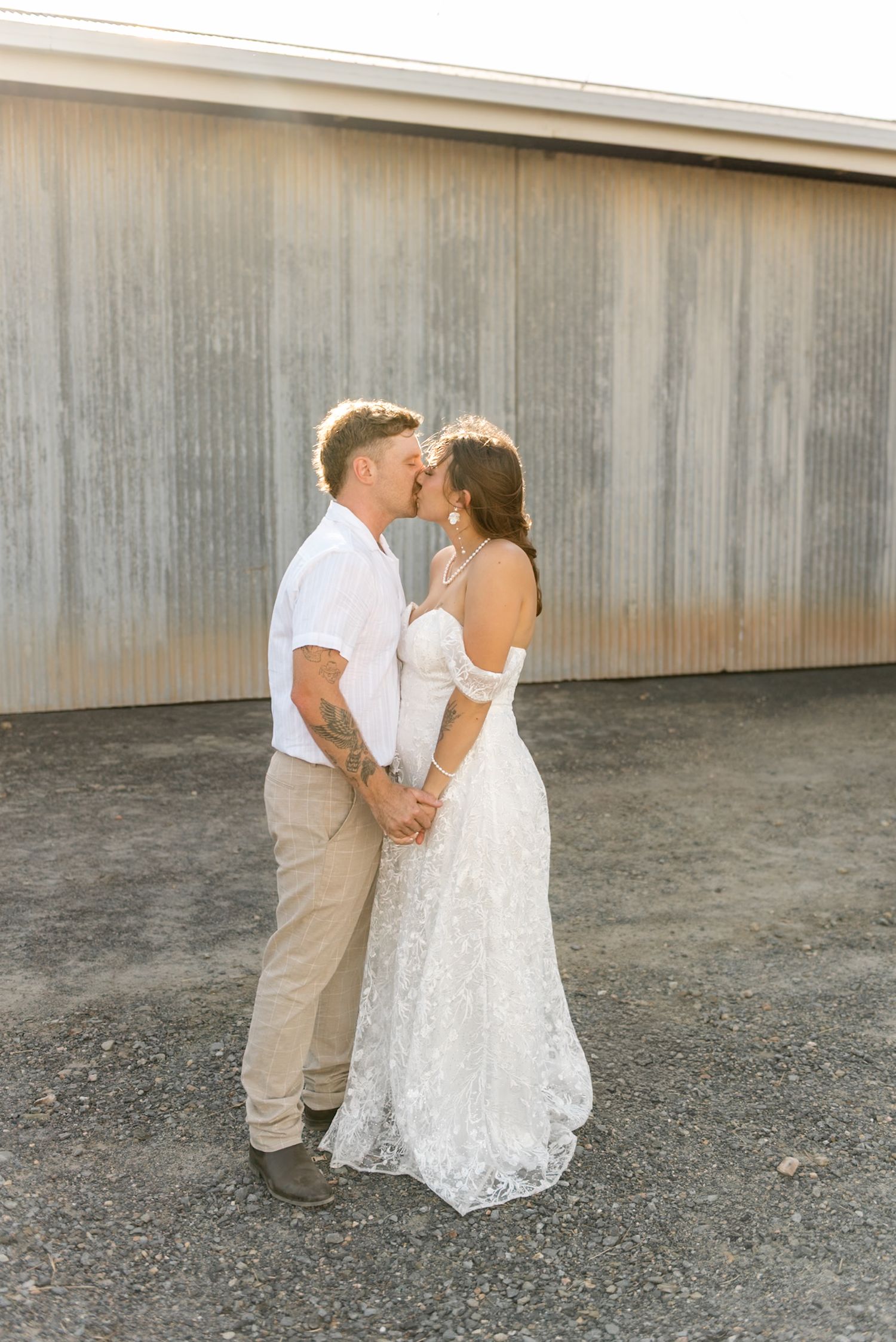 Newlyweds share a tender kiss holding hands in golden hour light before a long corrugated metal wall.