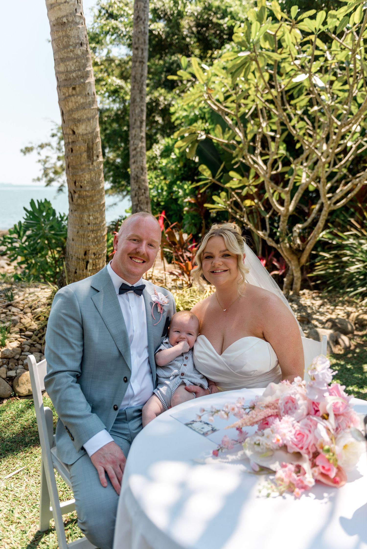 Smiling bride and groom in grey suit holding newborn baby at outdoor wedding signing table with pink flowers.
