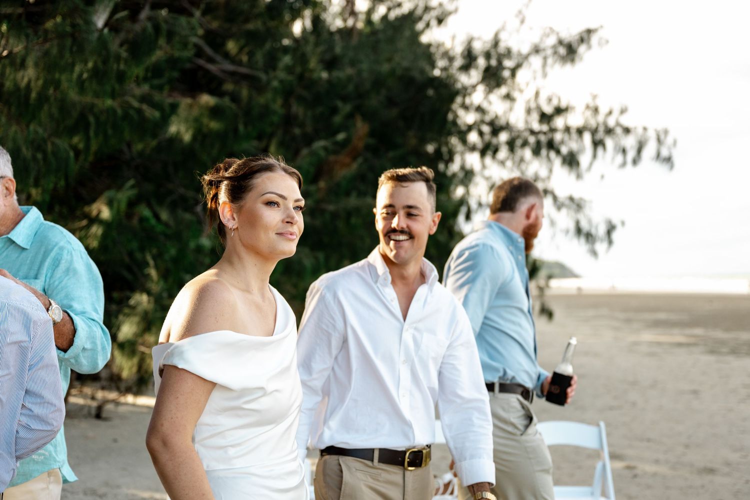 Smiling bride in white off-shoulder gown walks with groom at a tropical beach wedding at Four Mile Beach, Port Douglas.