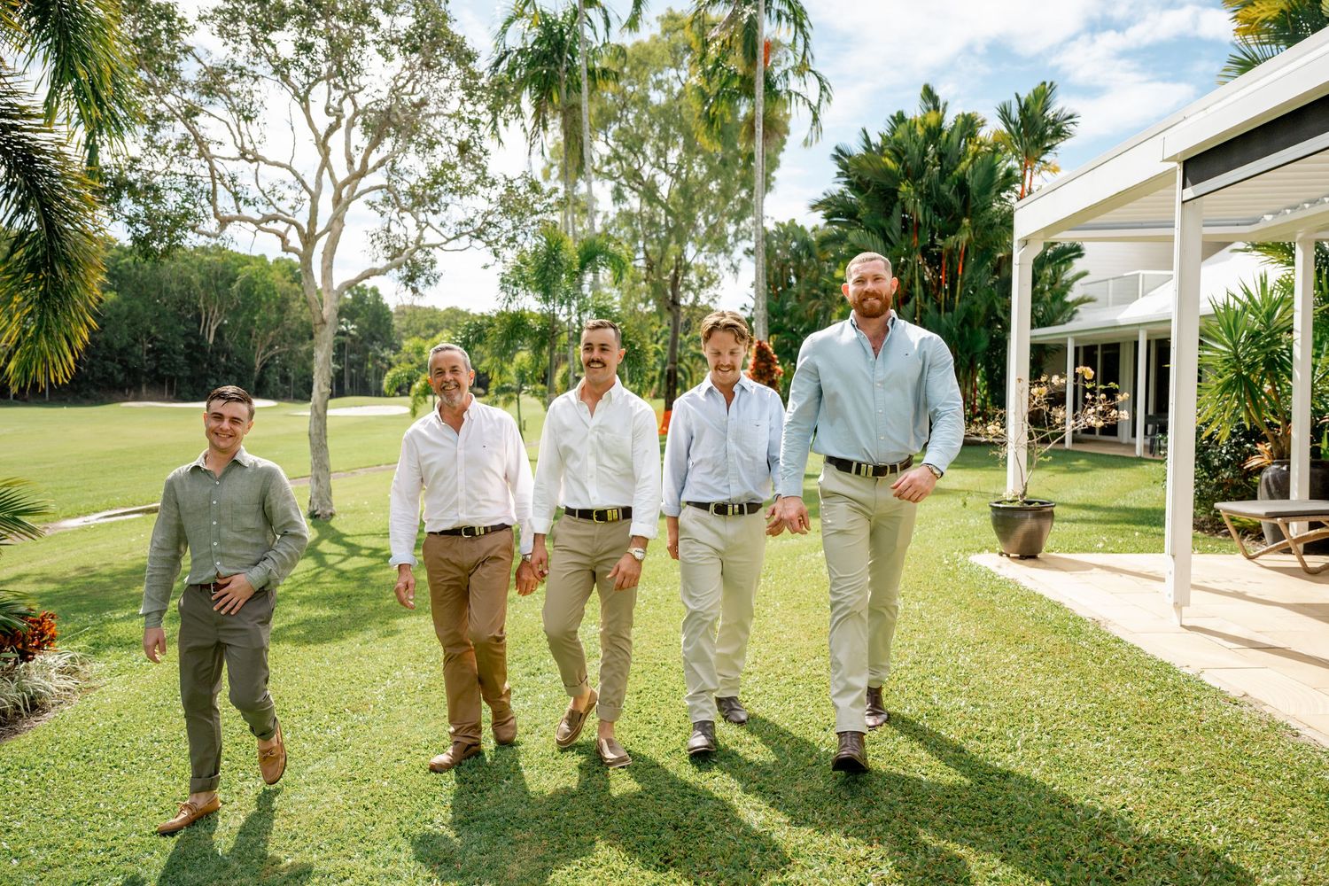 Six groomsmen in casual attire walking across lush green lawn surrounded by tropical palm trees.