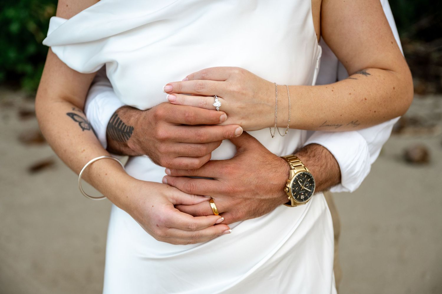 Close-up of bride and groom embracing, showing wedding rings, gold watch, and floral tattoo on their hands at Four Mile Beach, Port Douglas.