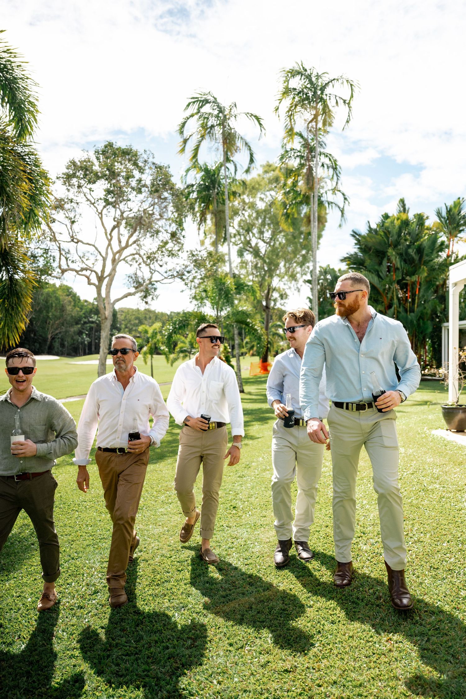 Group of groomsmen in smart casual attire strolling across lush green golf course on sunny day.