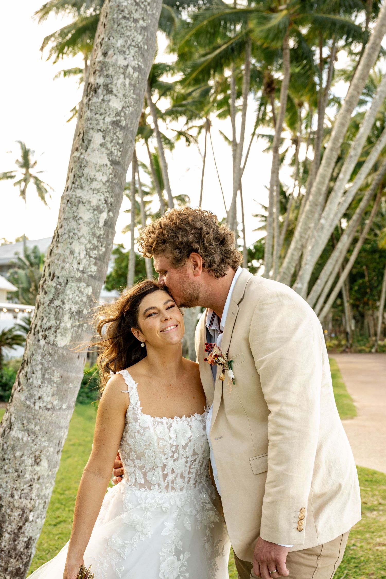 Groom tenderly kissing bride on the cheek as she smiles near a palm tree in a tropical garden.