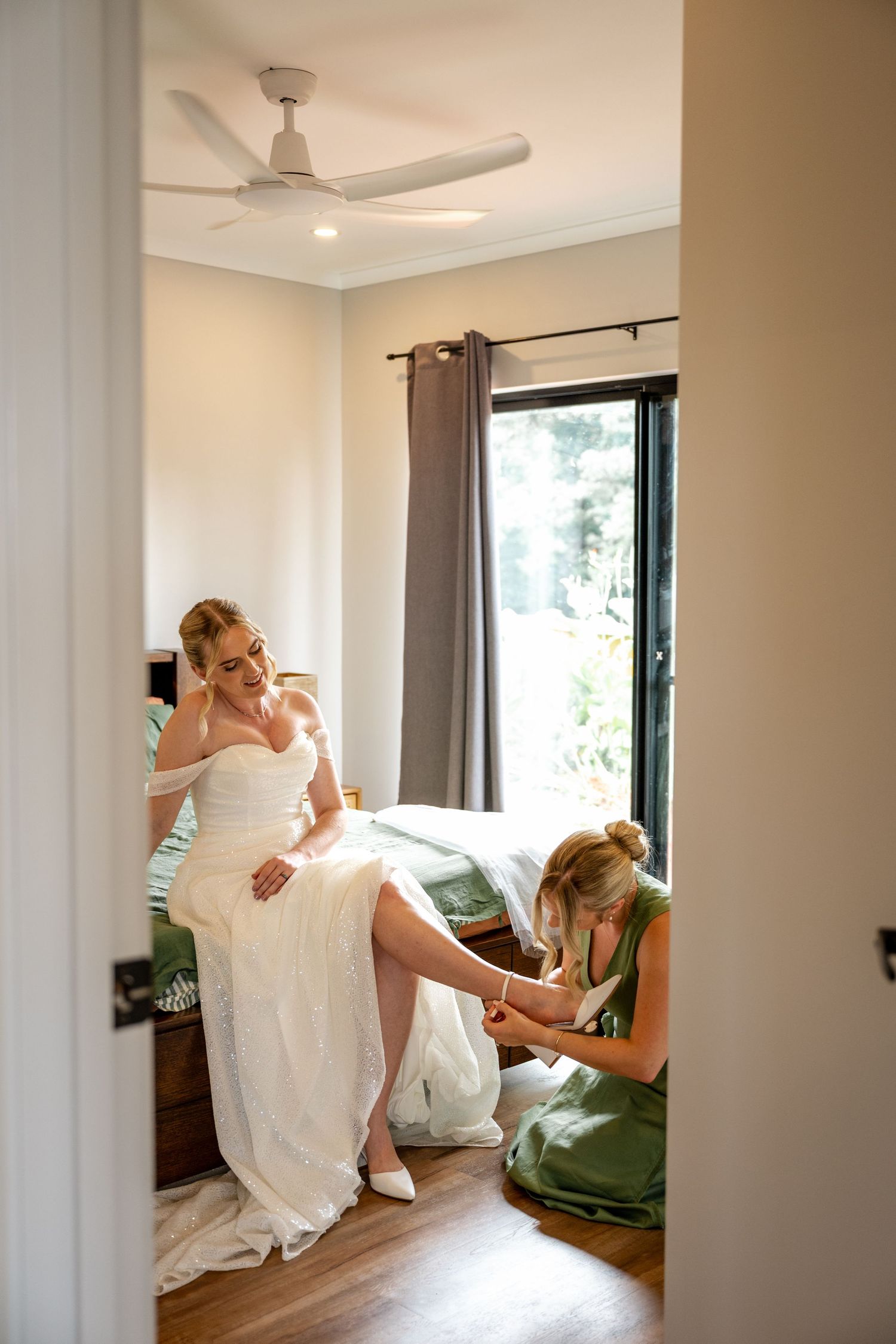 Bridesmaid in green dress helping blonde bride put on white heels in bedroom.