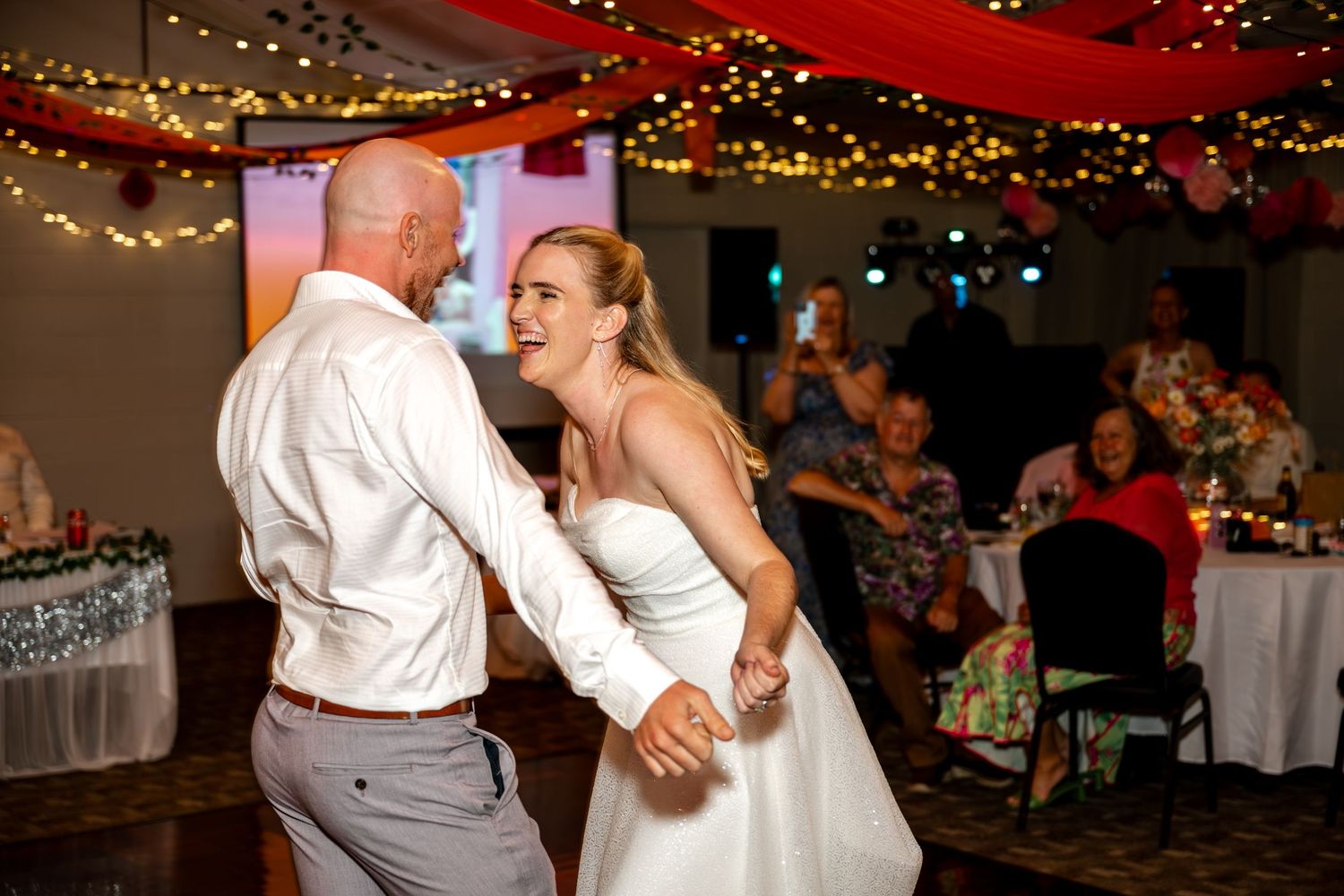 Smiling bride in white gown dances with groom in white shirt at a festive wedding reception.