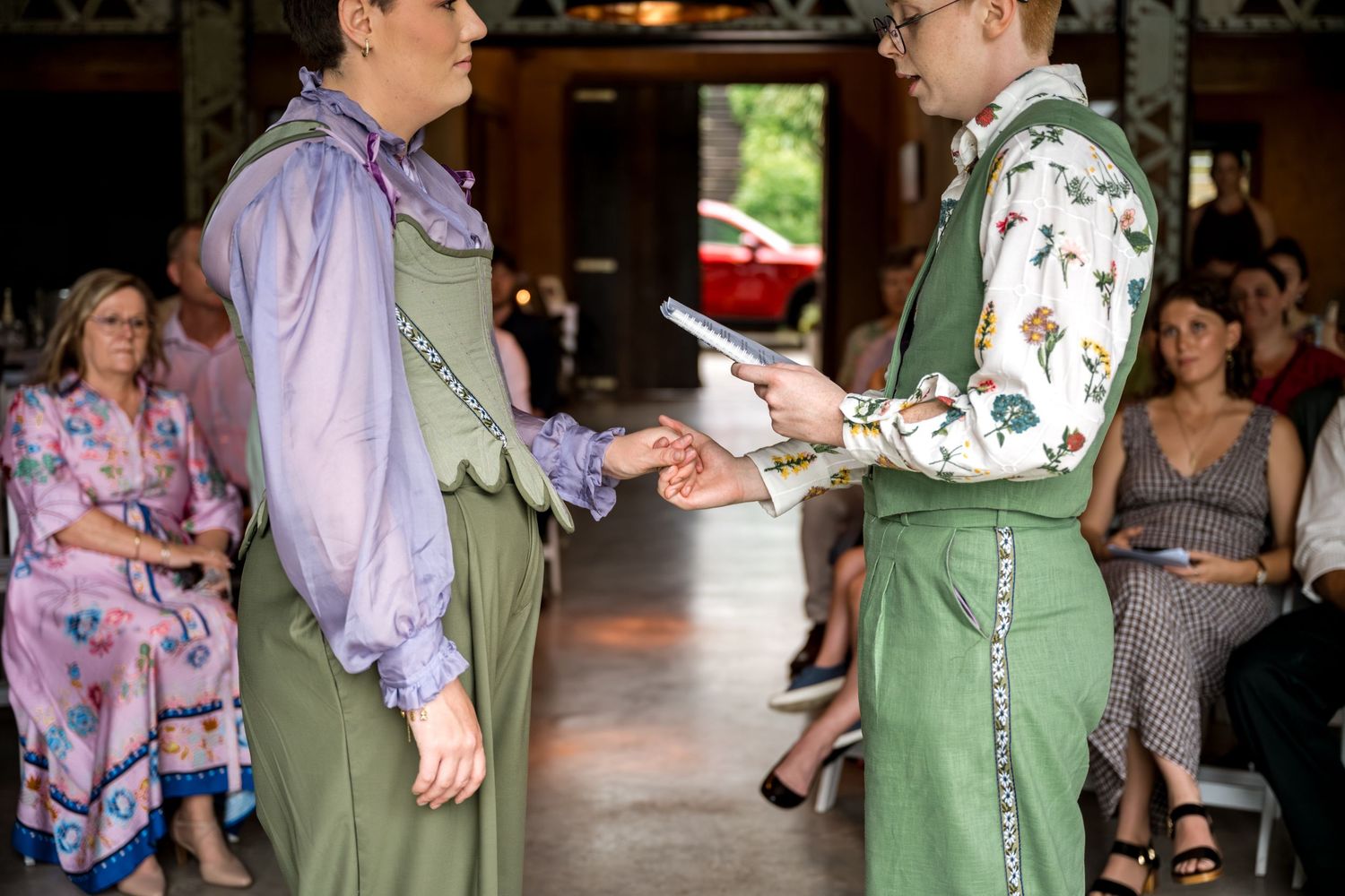 Two brides in matching green overalls exchange rings during intimate indoor wedding ceremony with guests seated behind.