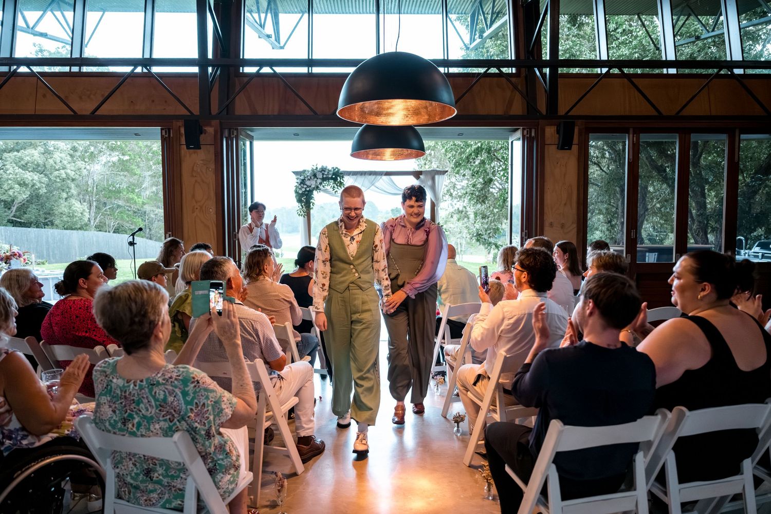 Two brides in green overalls and purple and floral blouses walk down aisle after wedding ceremony.