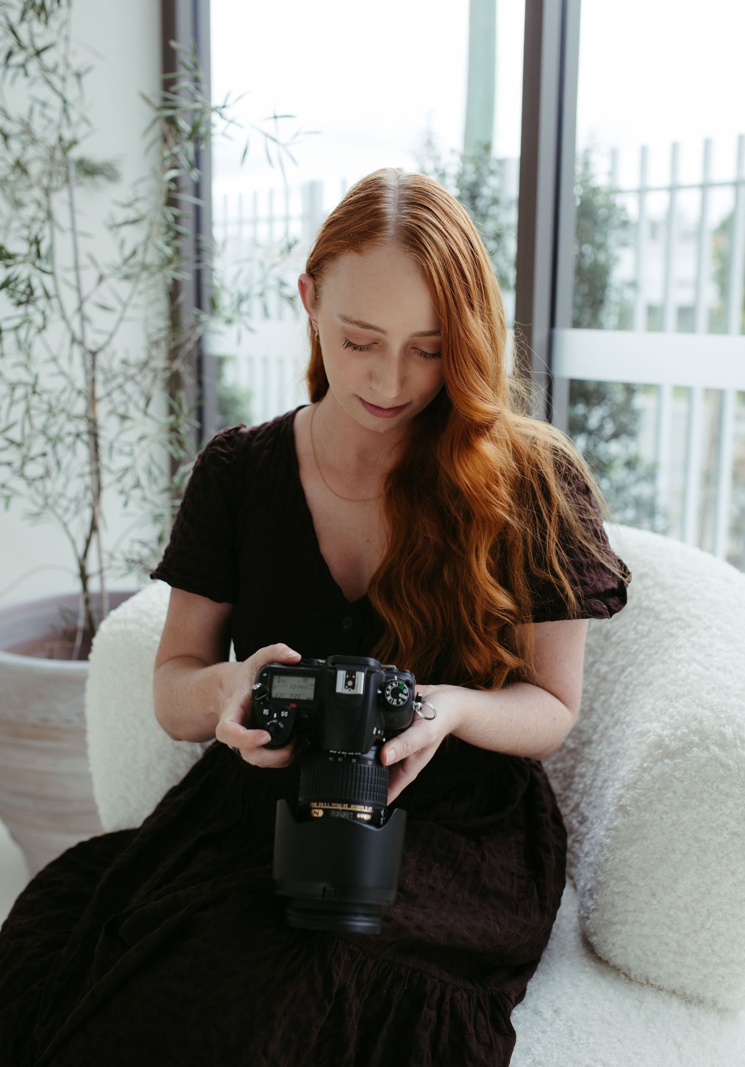 Redhead female photographer in black dress reviewing images on DSLR camera indoors.