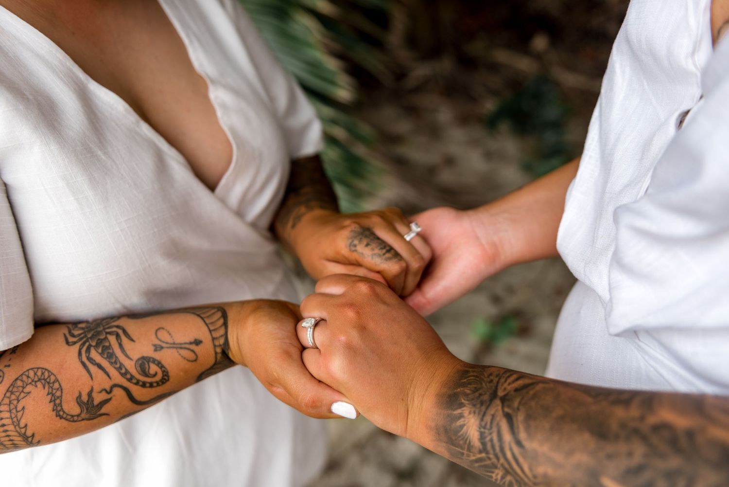 Close-up of two brides holding hands with intricate tattoos visible on their arms, wearing wedding rings.