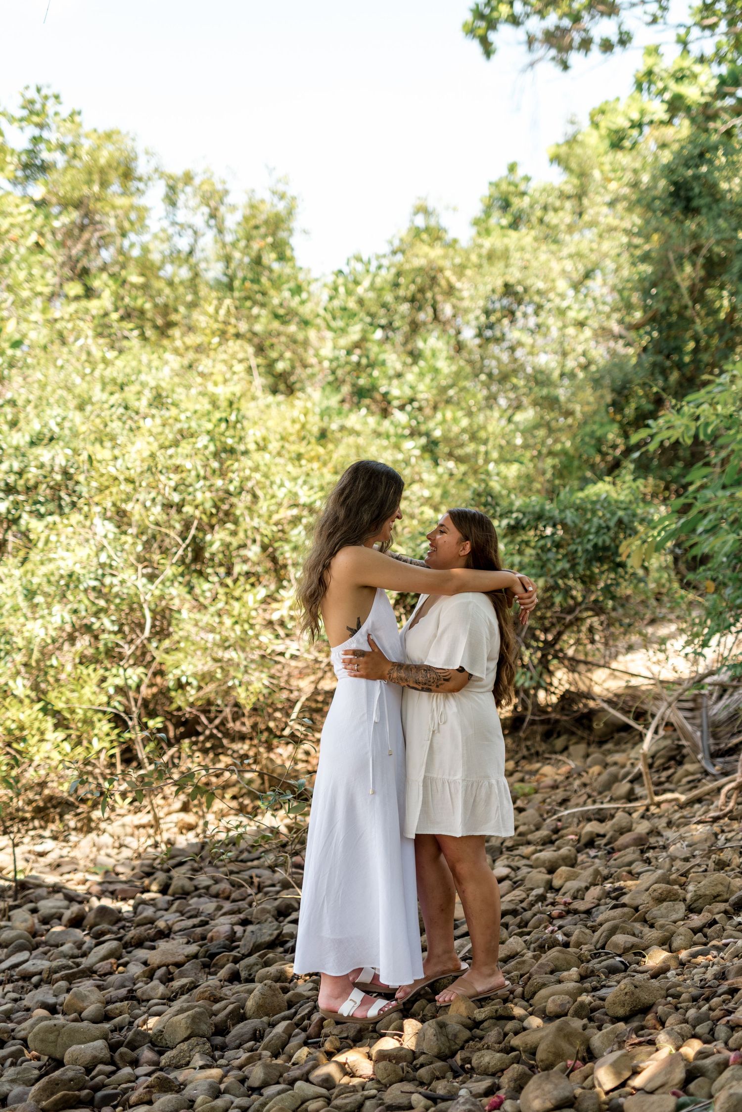 Two women in white dresses share a close embrace on a rocky riverbed surrounded by lush green vegetation.