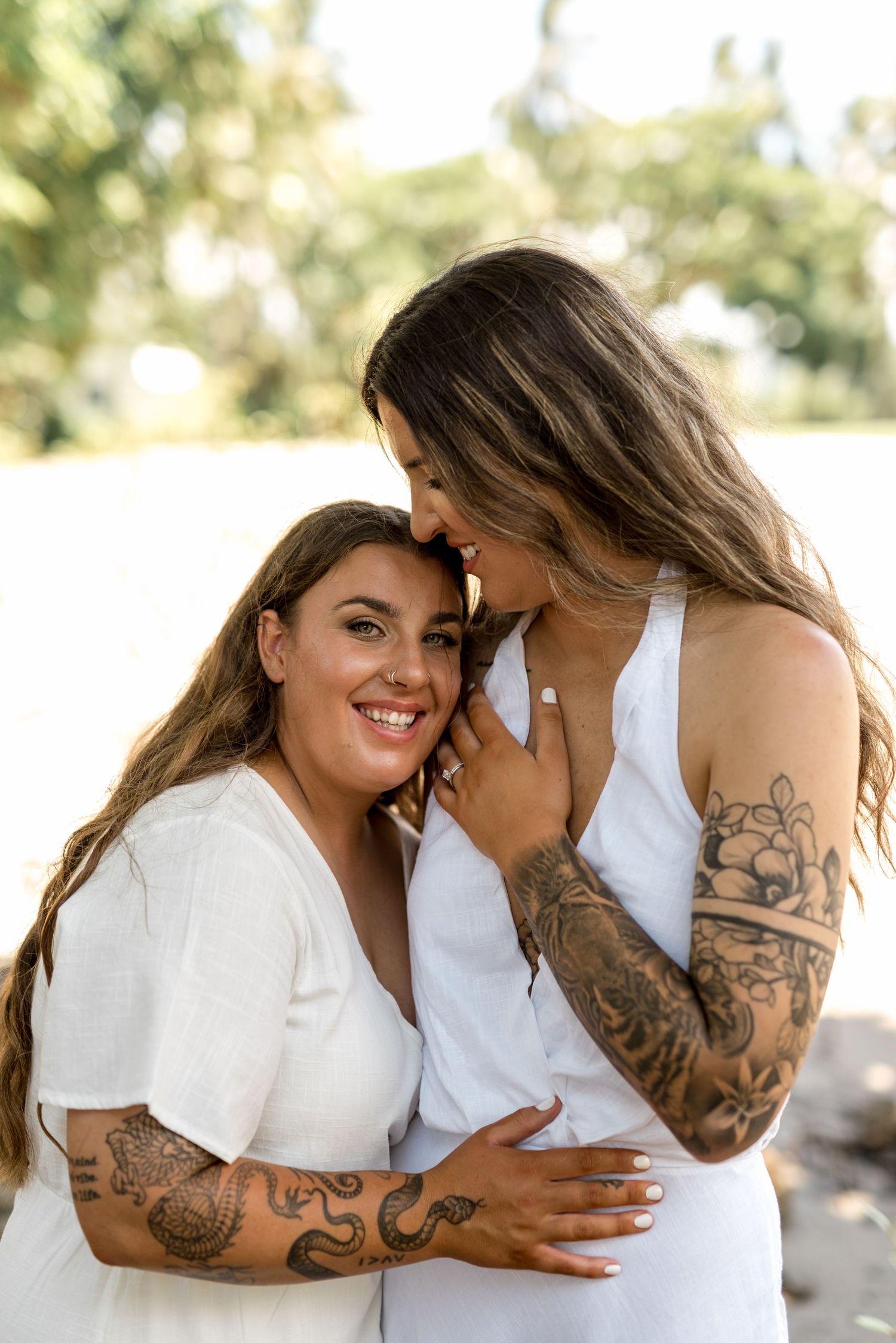 Two tattooed women in white dresses share an intimate moment outdoors, one smiling at the camera in warm sunlight.
