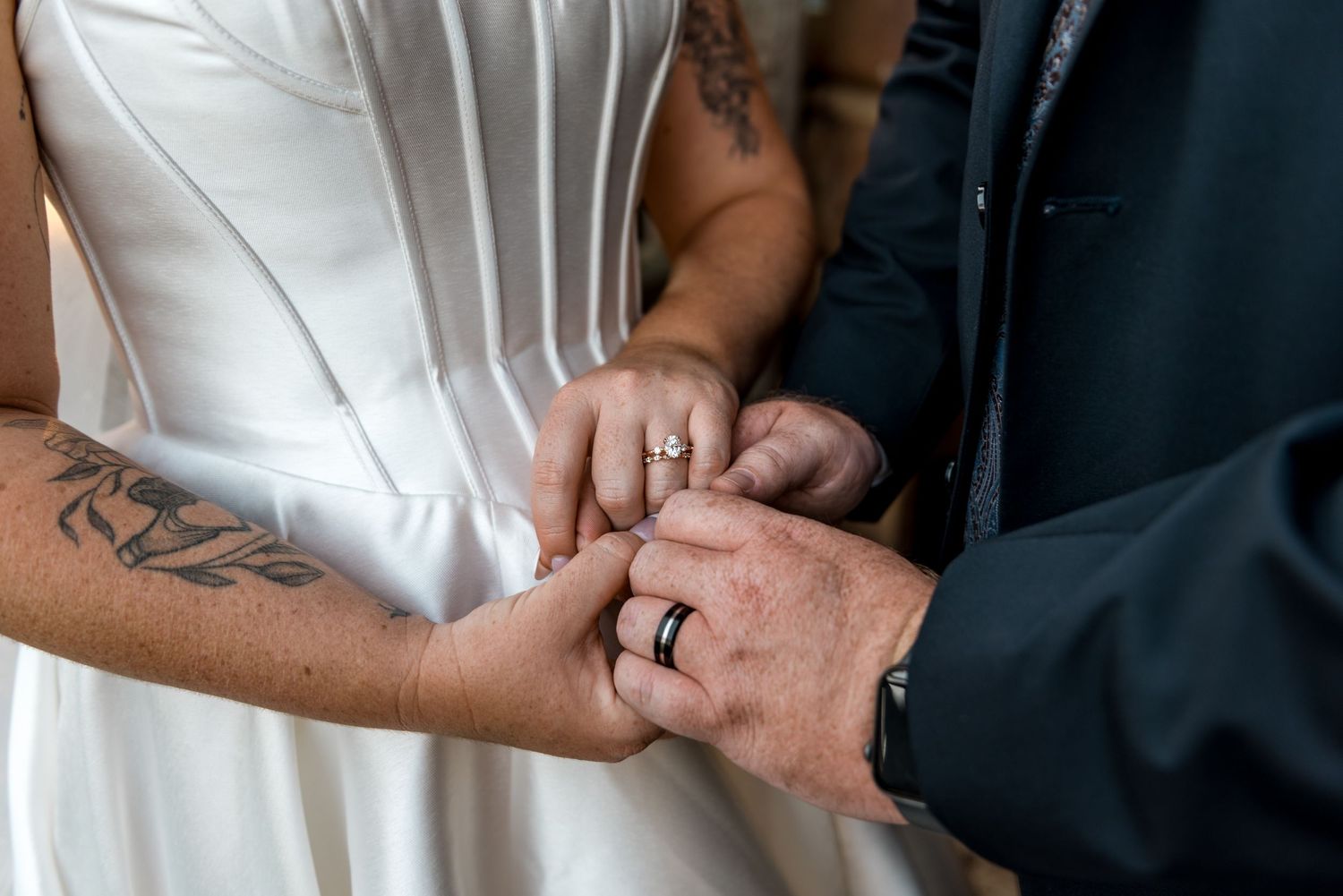 Close-up of bride and groom exchanging rings showing tattoos and wedding bands.