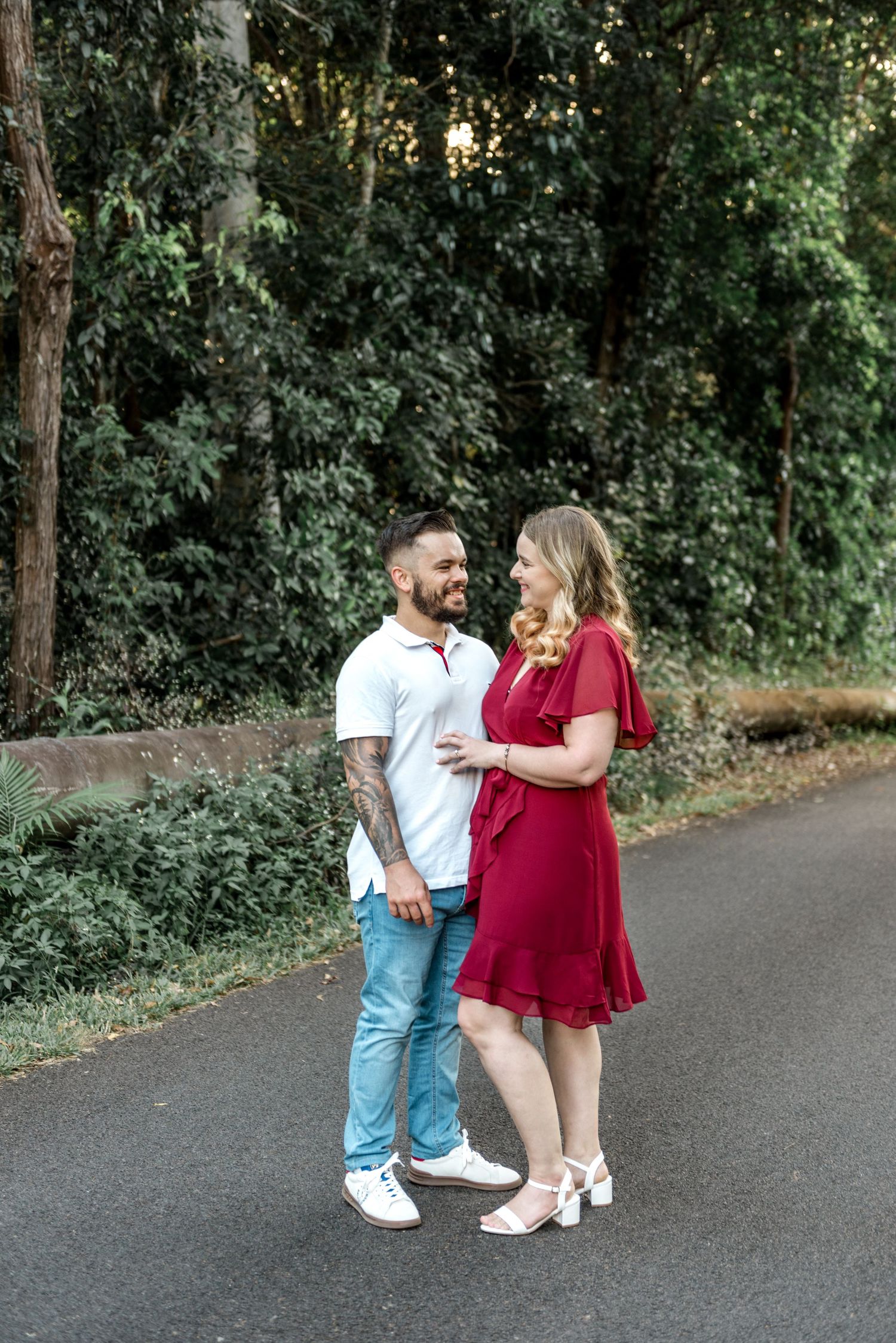 Couple in coordinating outfits sharing a loving gaze while standing on a tree-lined road in warm light.