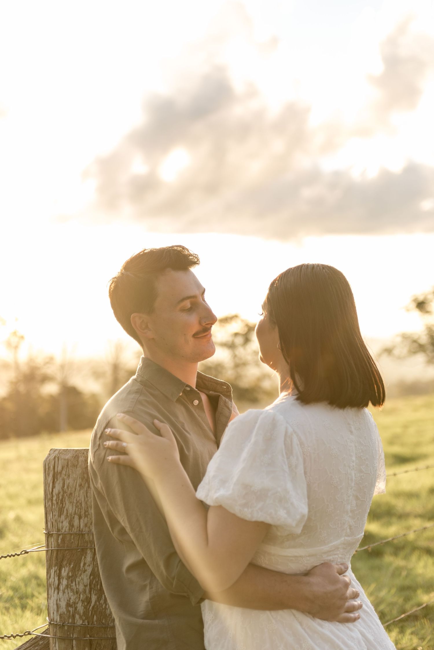 Couple gazing into each others eyes at golden hour by a rustic fence in a green field.