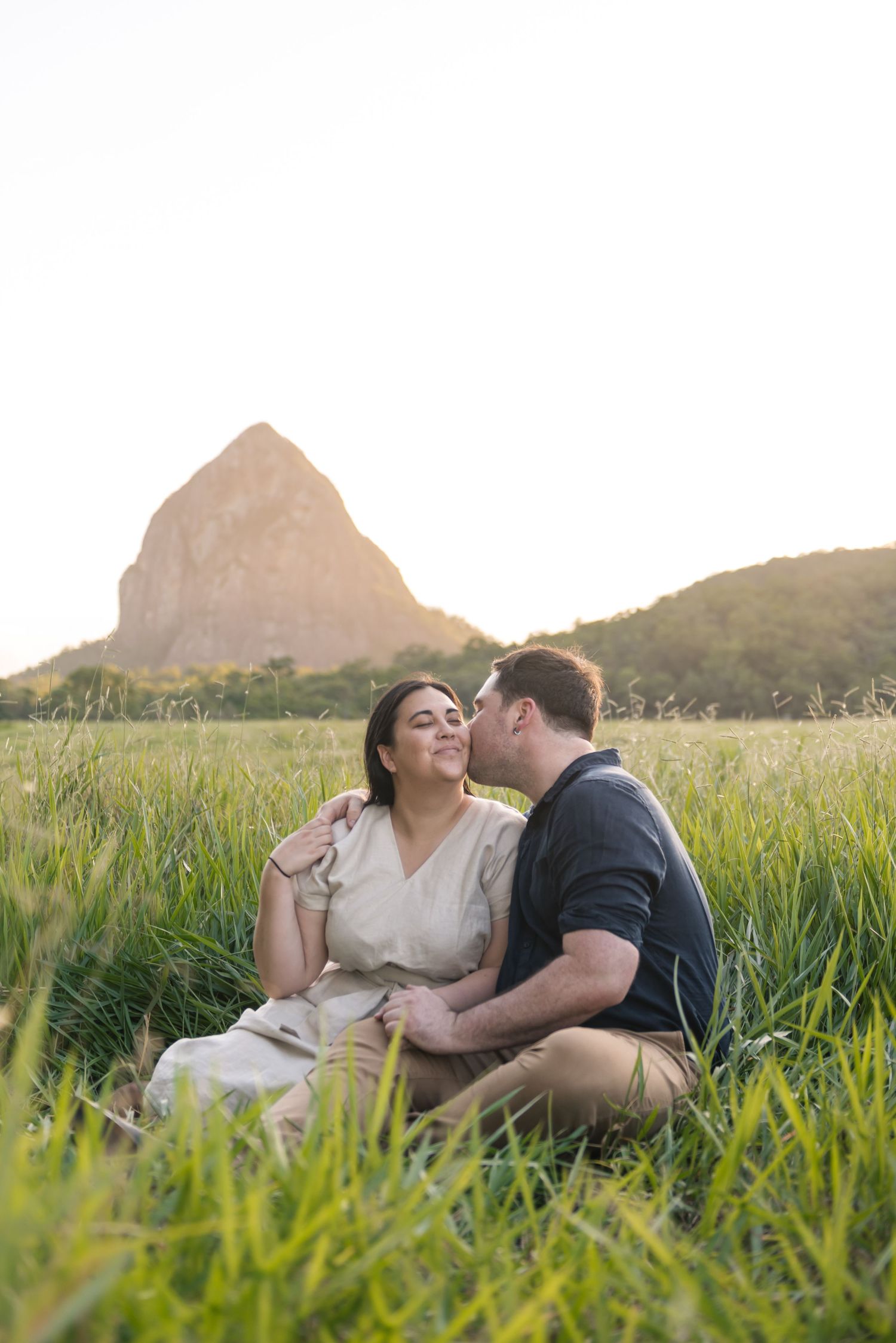 Man kissing woman on cheek while sitting in lush green field at sunset.