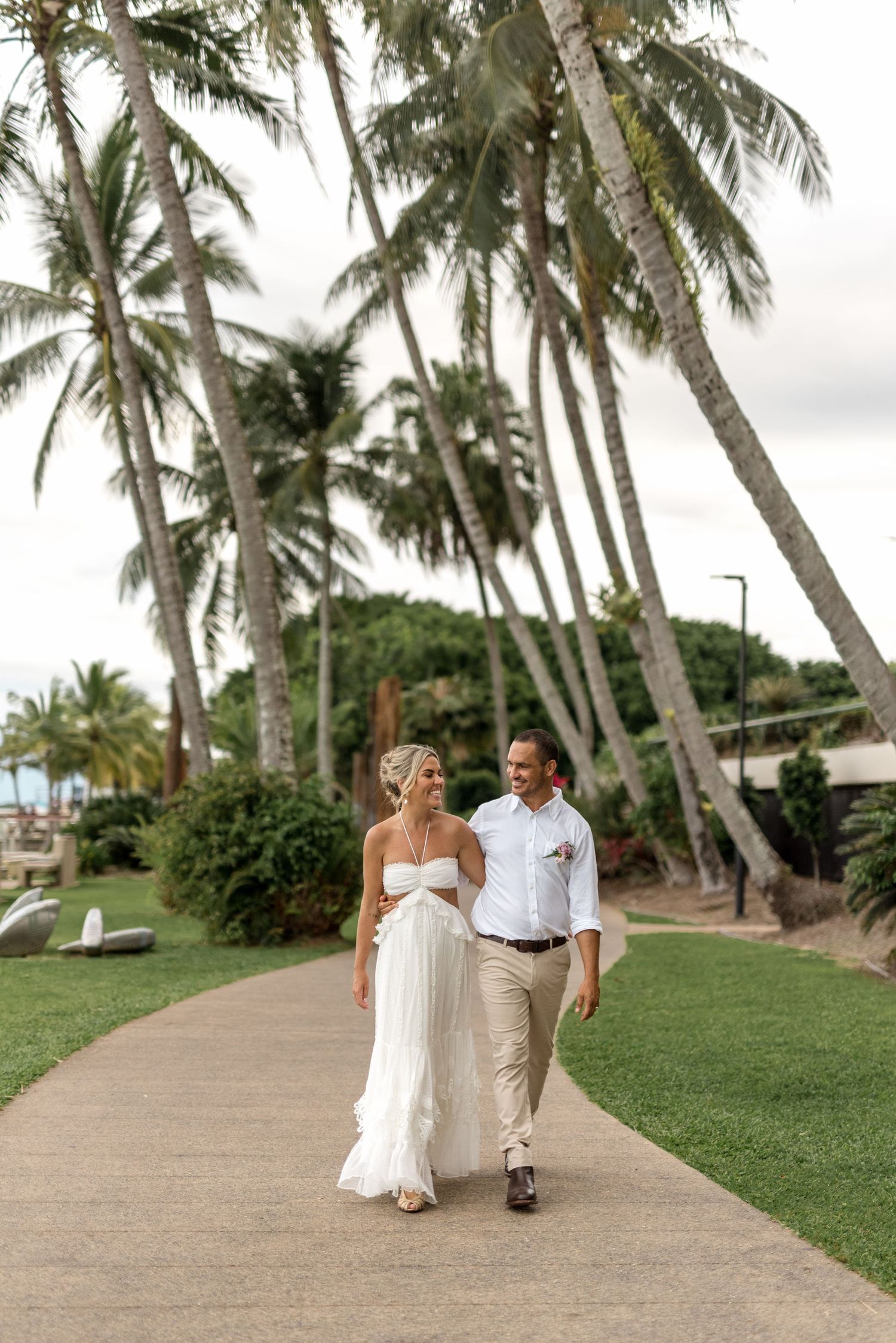 Bride and groom walking hand in hand along a tropical garden path lined with tall swaying palm trees.