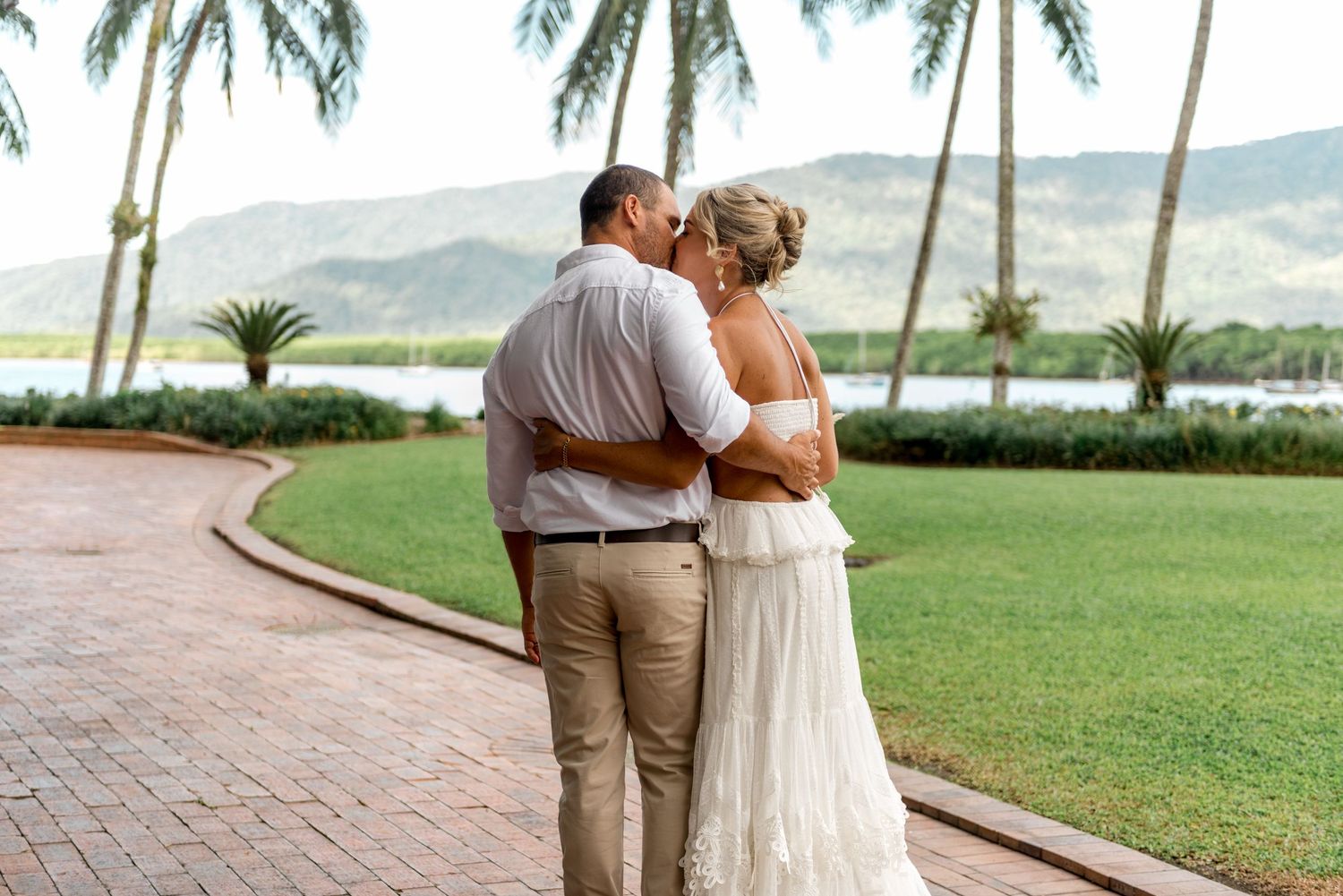 Bride and groom share a romantic kiss in a lush tropical garden with palm trees and mountains.