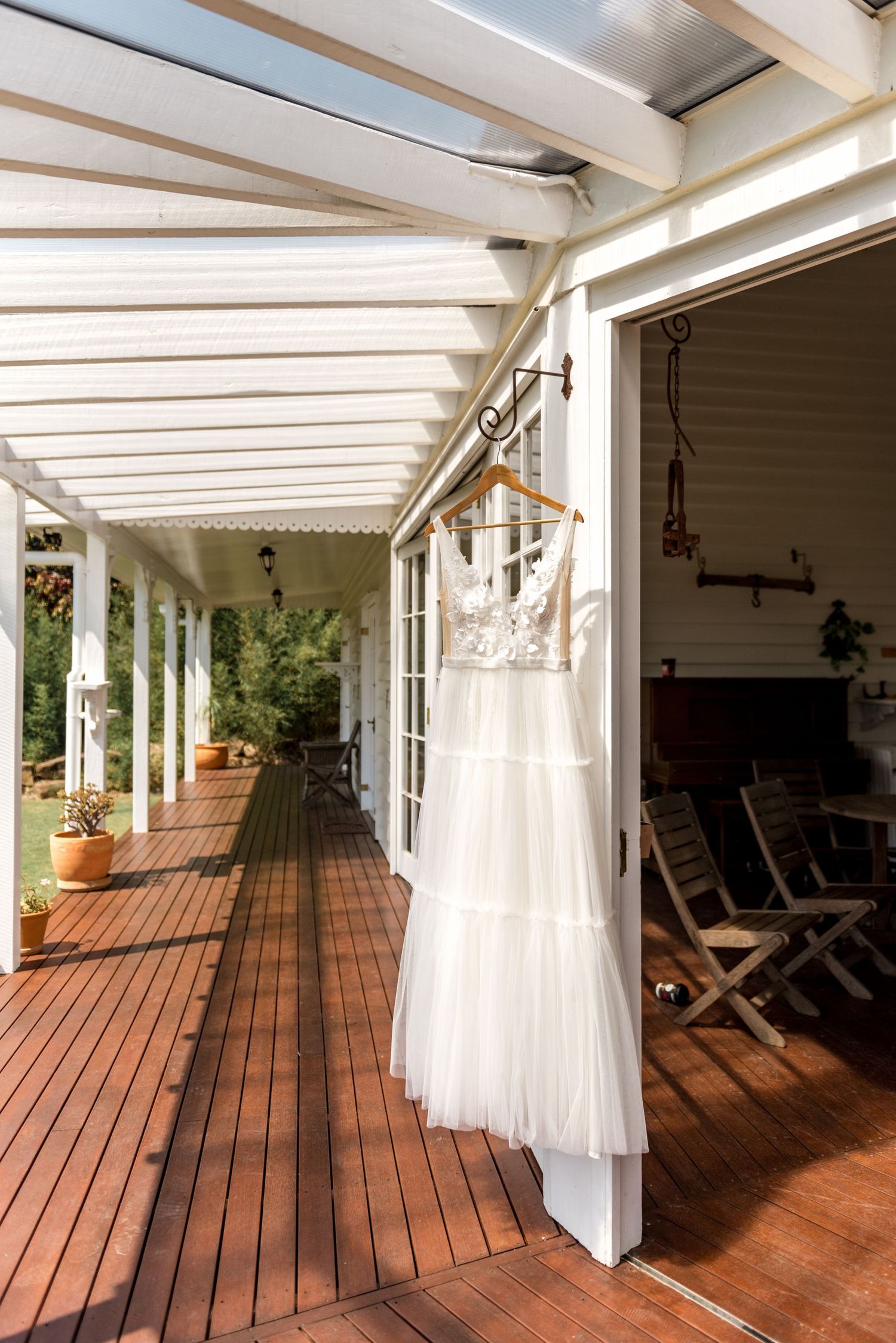 White lace wedding dress hanging on a hanger on a sunlit farmhouse verandah.