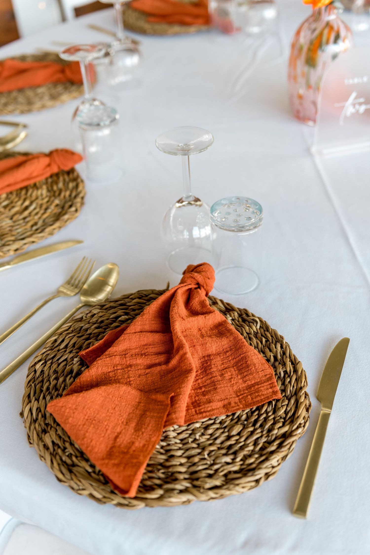 Elegant wedding table setting with orange linen napkin, woven charger plate and gold cutlery.