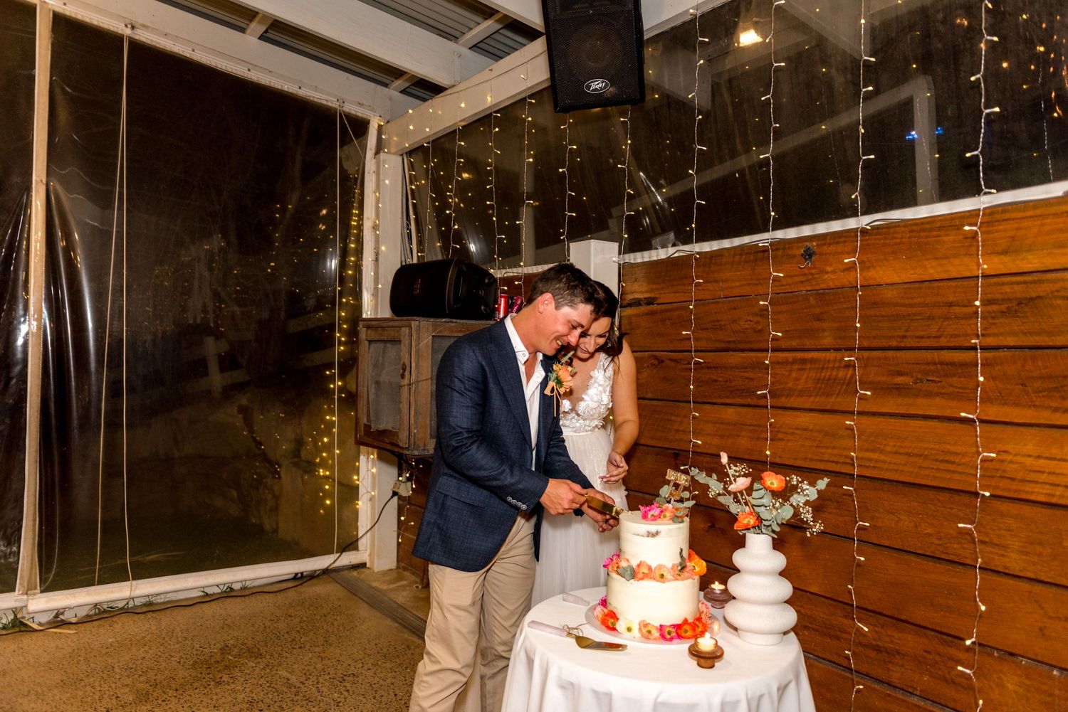 Bride and groom cut their floral-decorated wedding cake together in a warmly lit rustic barn reception venue.