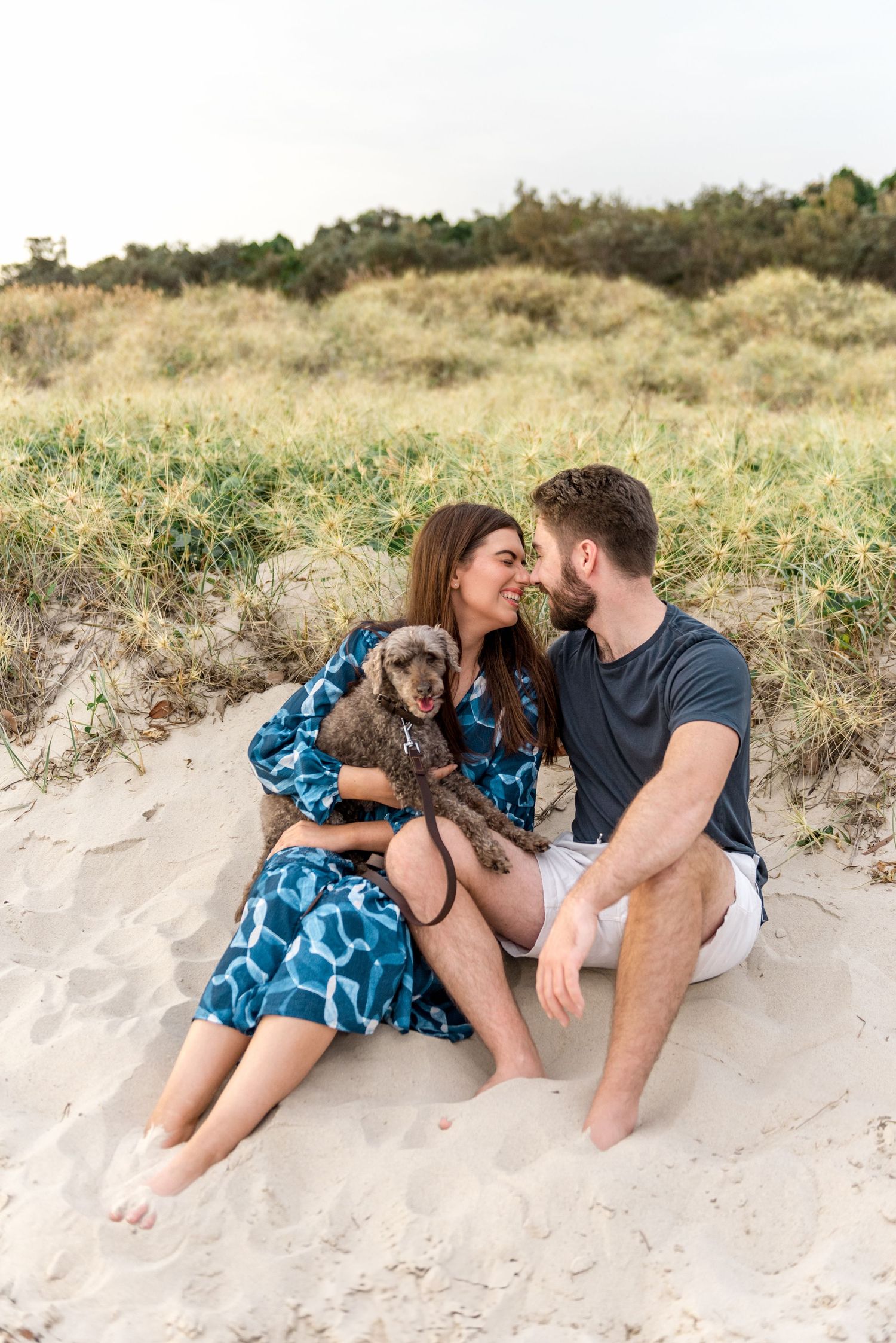 Couple sitting close on sandy beach dunes about to kiss while holding gray fluffy dog.