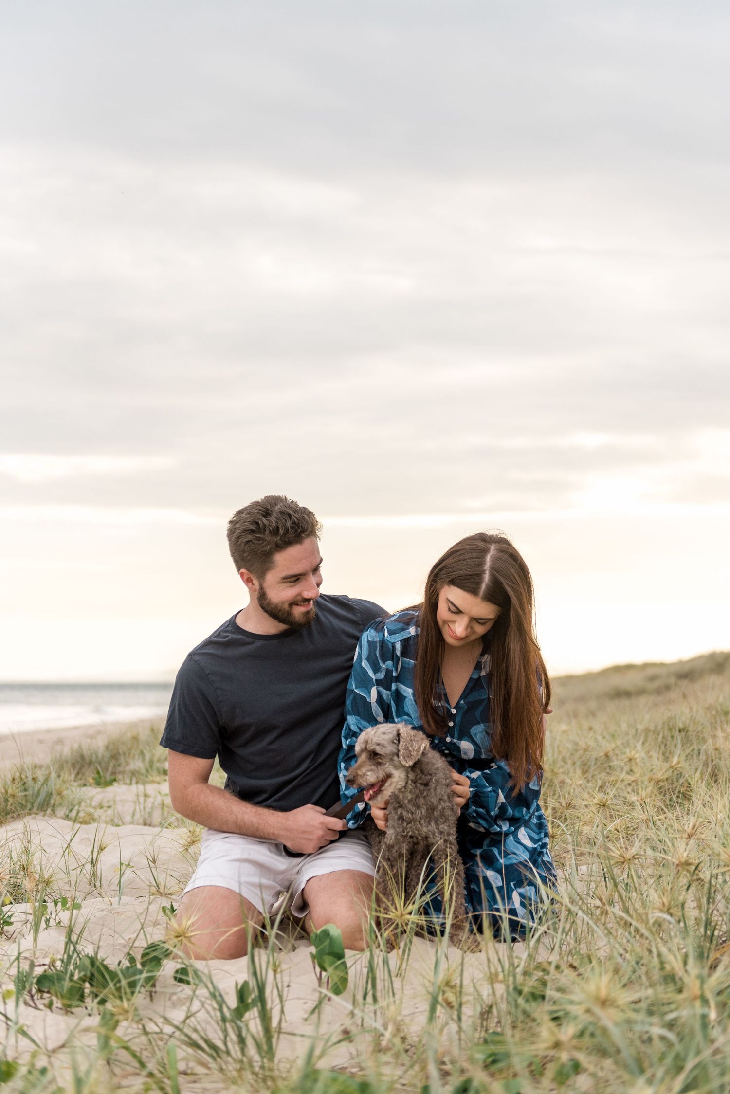 Couple sitting in coastal sand dunes with their small grey dog under a soft cloudy sky.