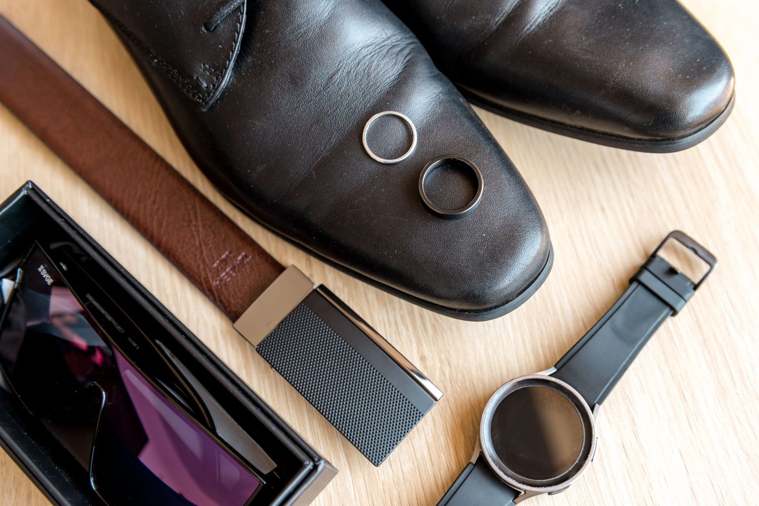Close-up flat lay of grooms accessories including black shoes, wedding rings, watch, belt, and sunglasses.