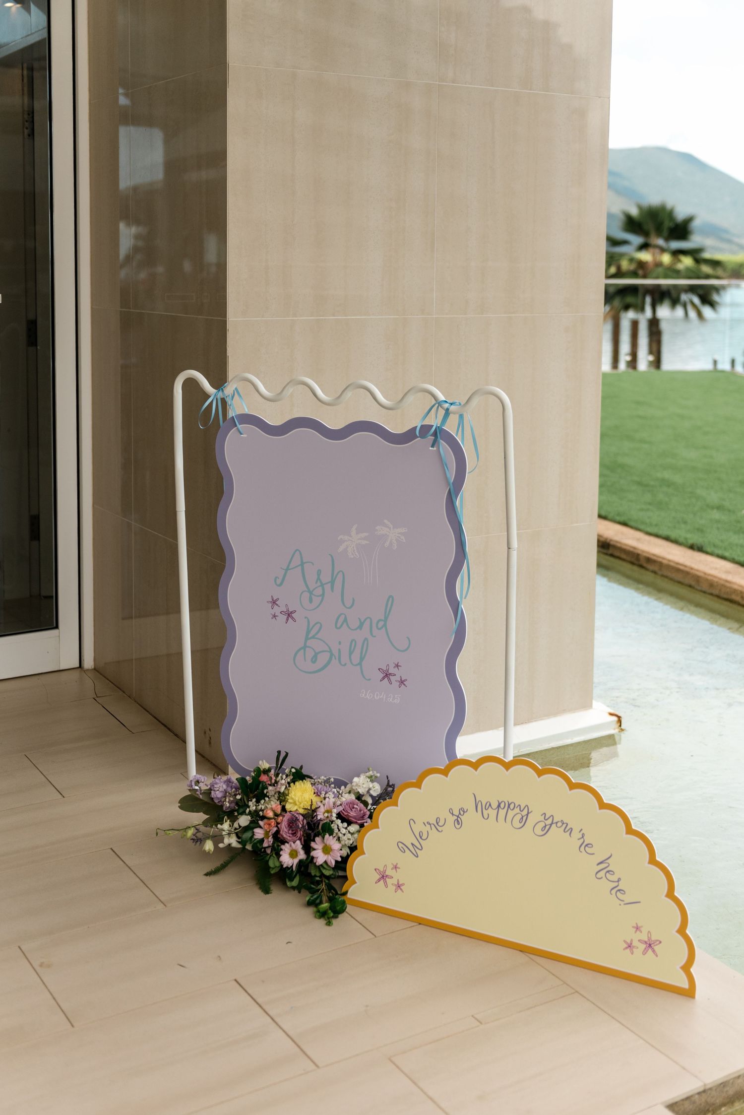 Colorful wedding welcome signs for Ash and Bill displayed on white stand with floral arrangement.