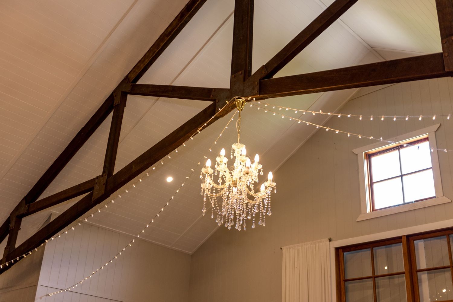 Rustic barn wedding venue interior featuring exposed timber beams, crystal chandelier, and delicate fairy string lights.