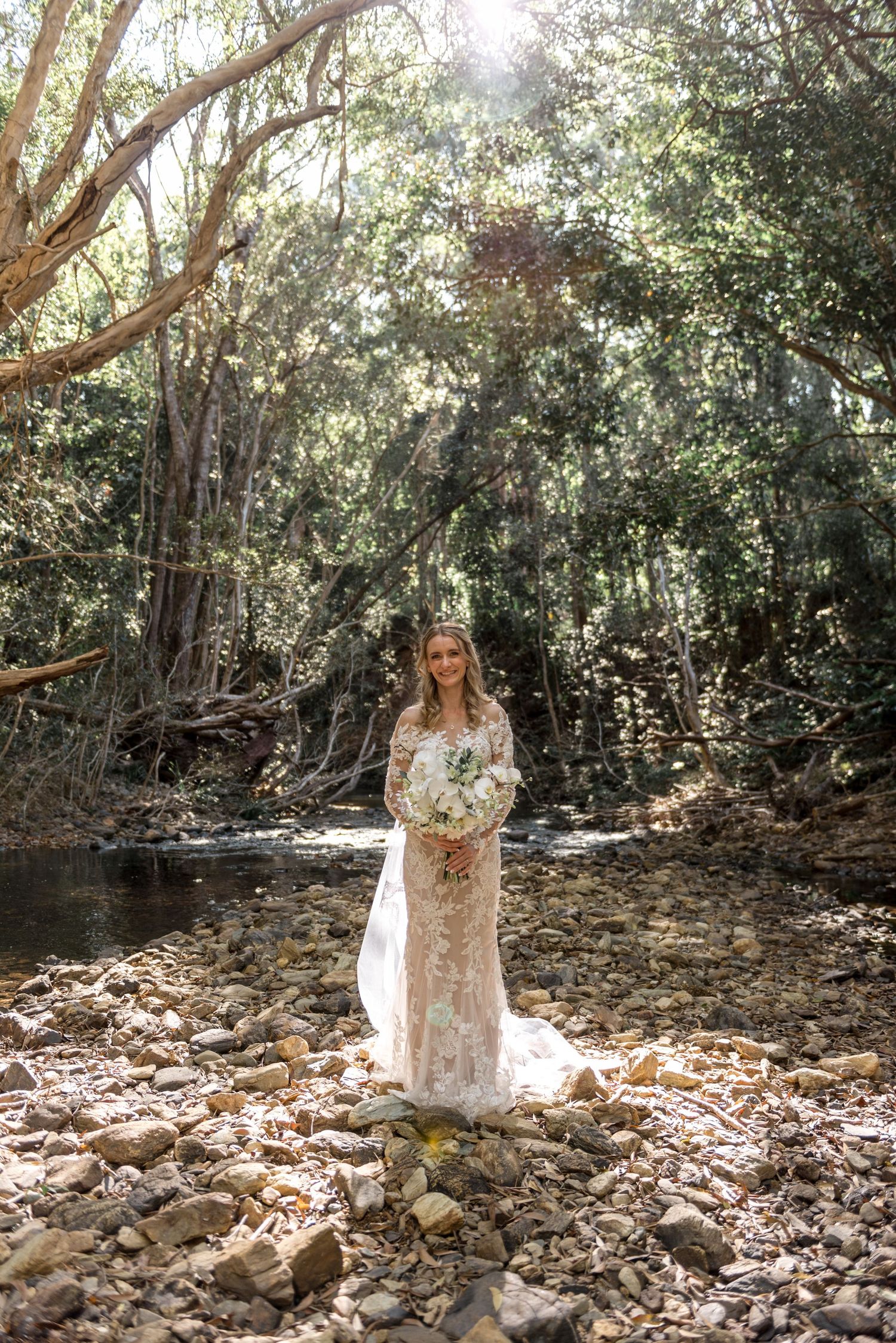 Bride in lace gown holds white bouquet standing on rocky creek bed in forest.