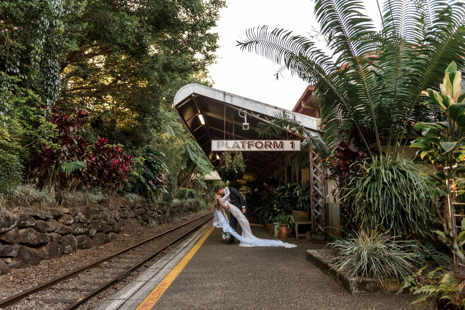 Bride and groom kiss under a rustic Platform 1 sign surrounded by tropical greenery at a train station.