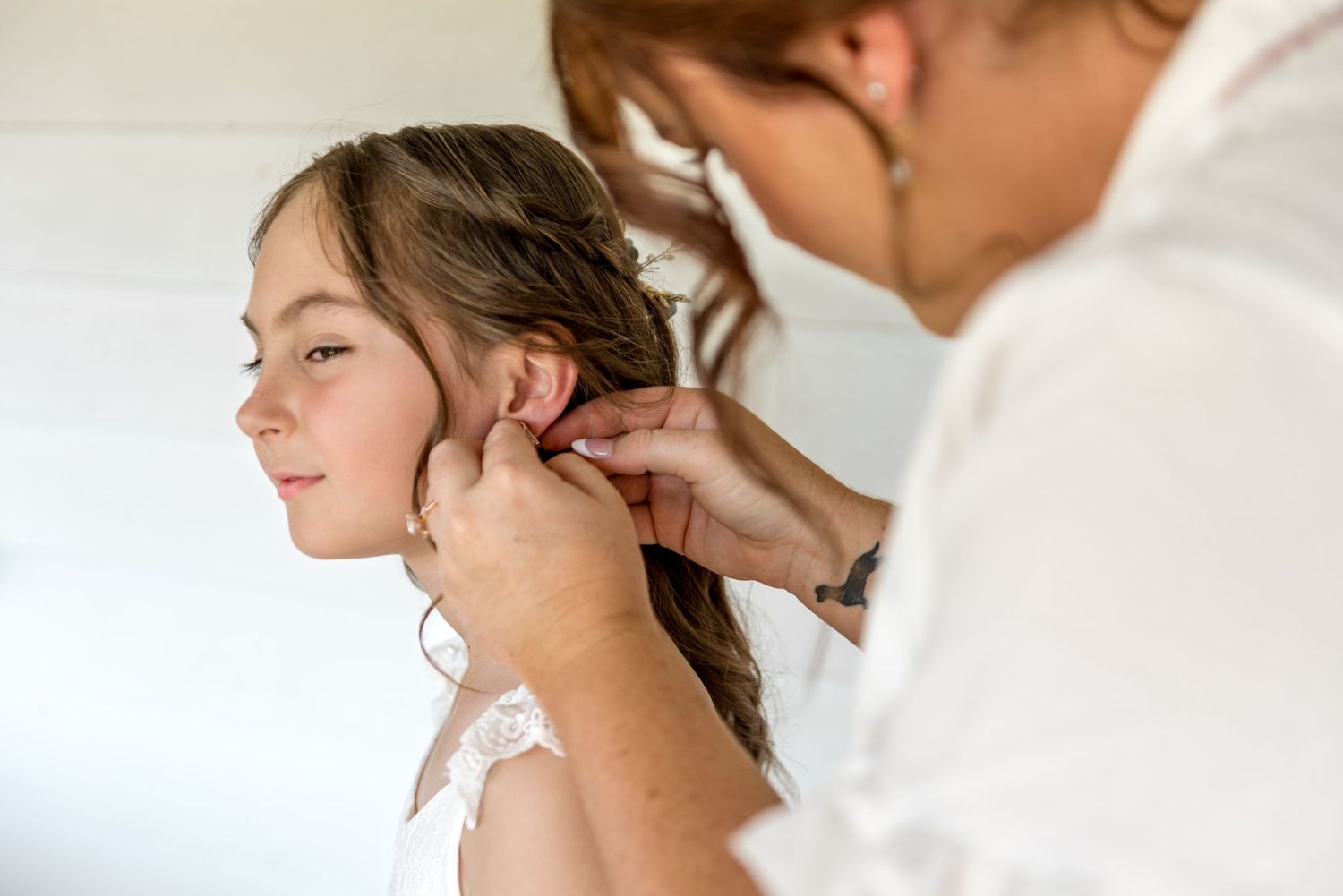 Woman carefully places earring on young flower girl with braided hair during wedding preparations.