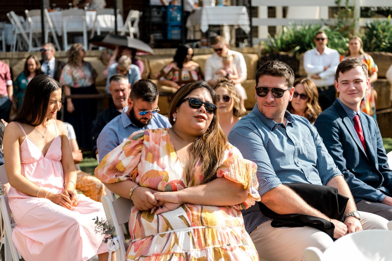 Smiling wedding guests seated in white chairs enjoying an outdoor ceremony on a sunny day.