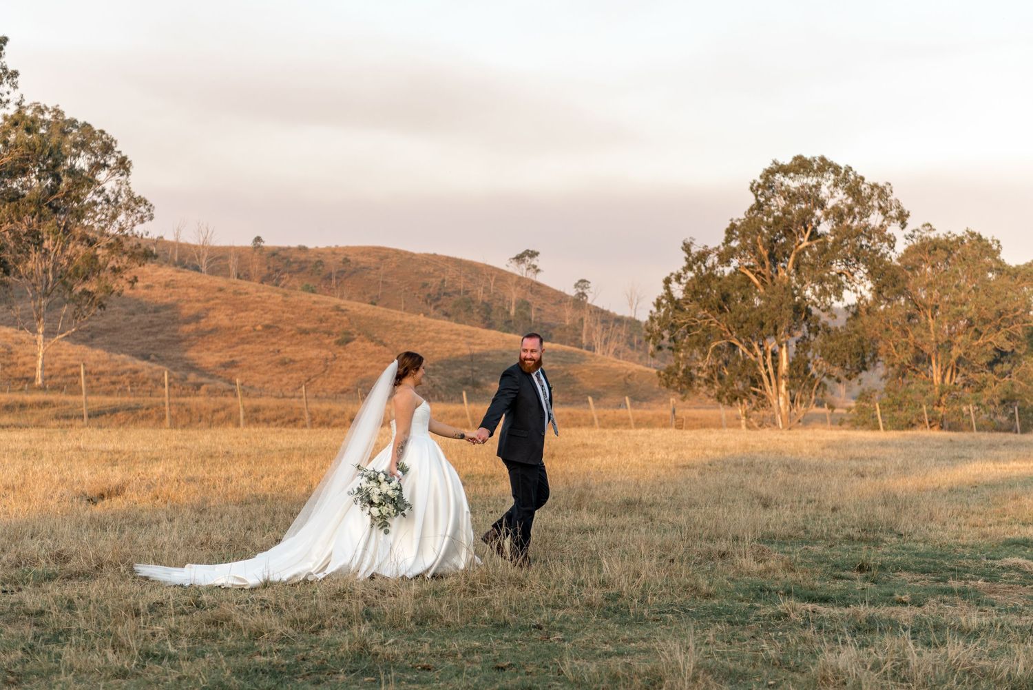 Bride and groom walking hand in hand through golden grass field at dusk with scenic hills.