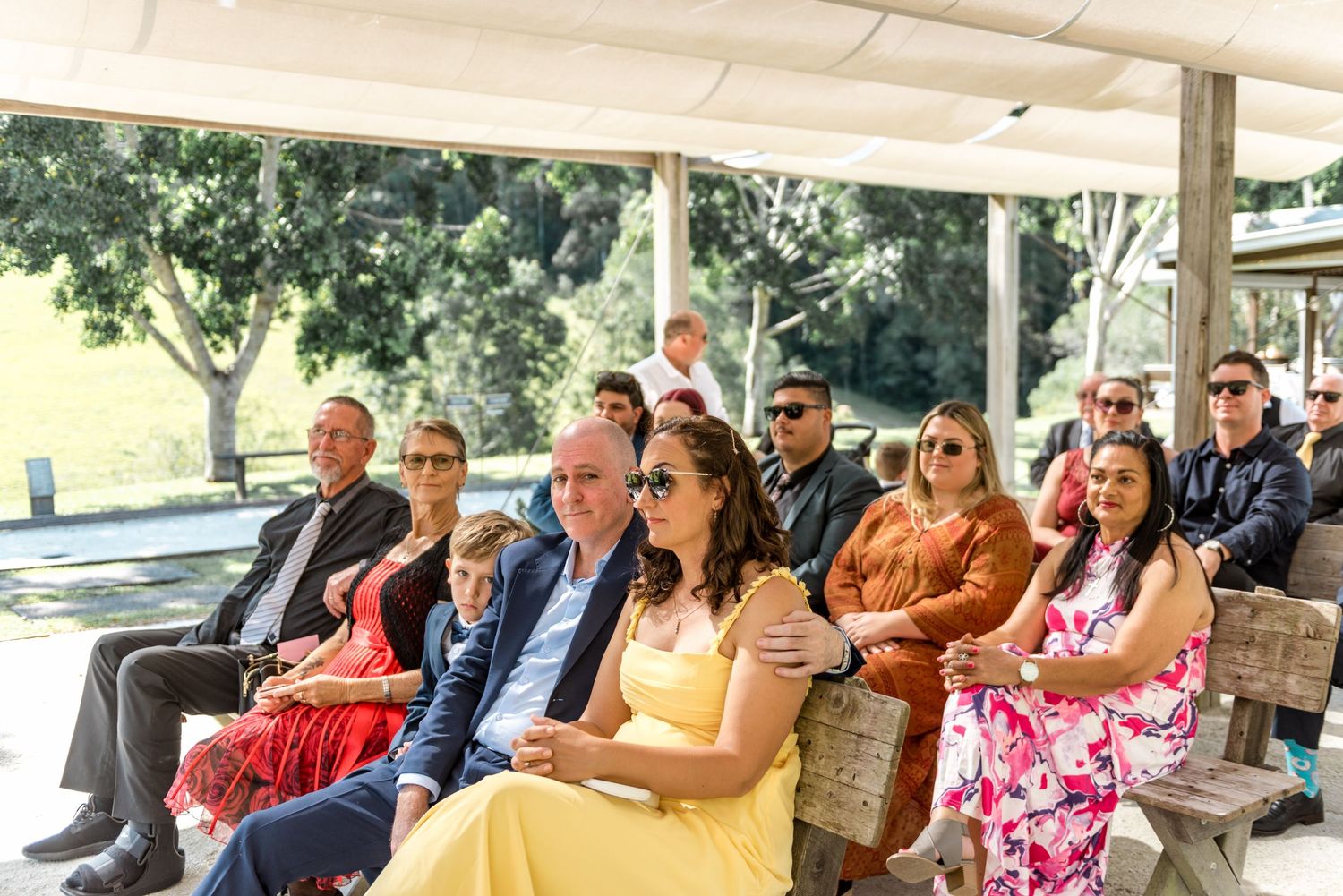 Wedding guests seated outdoors on wooden benches enjoying a sunny outdoor wedding ceremony.