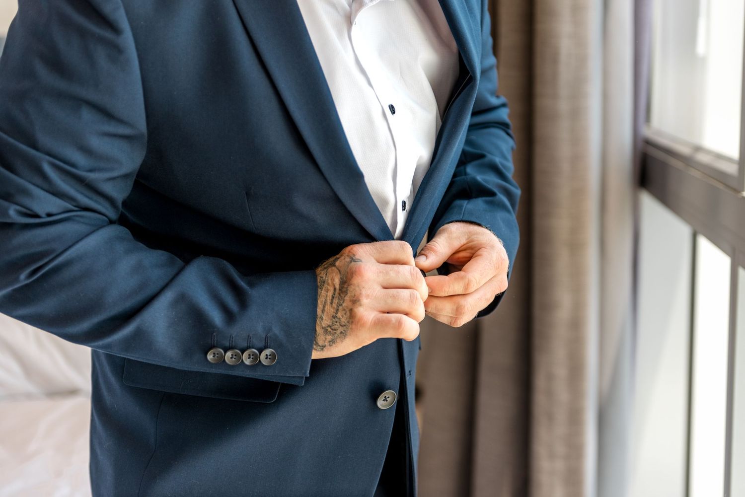 Groom in navy suit buttoning jacket, revealing tattooed hands near a bright window.