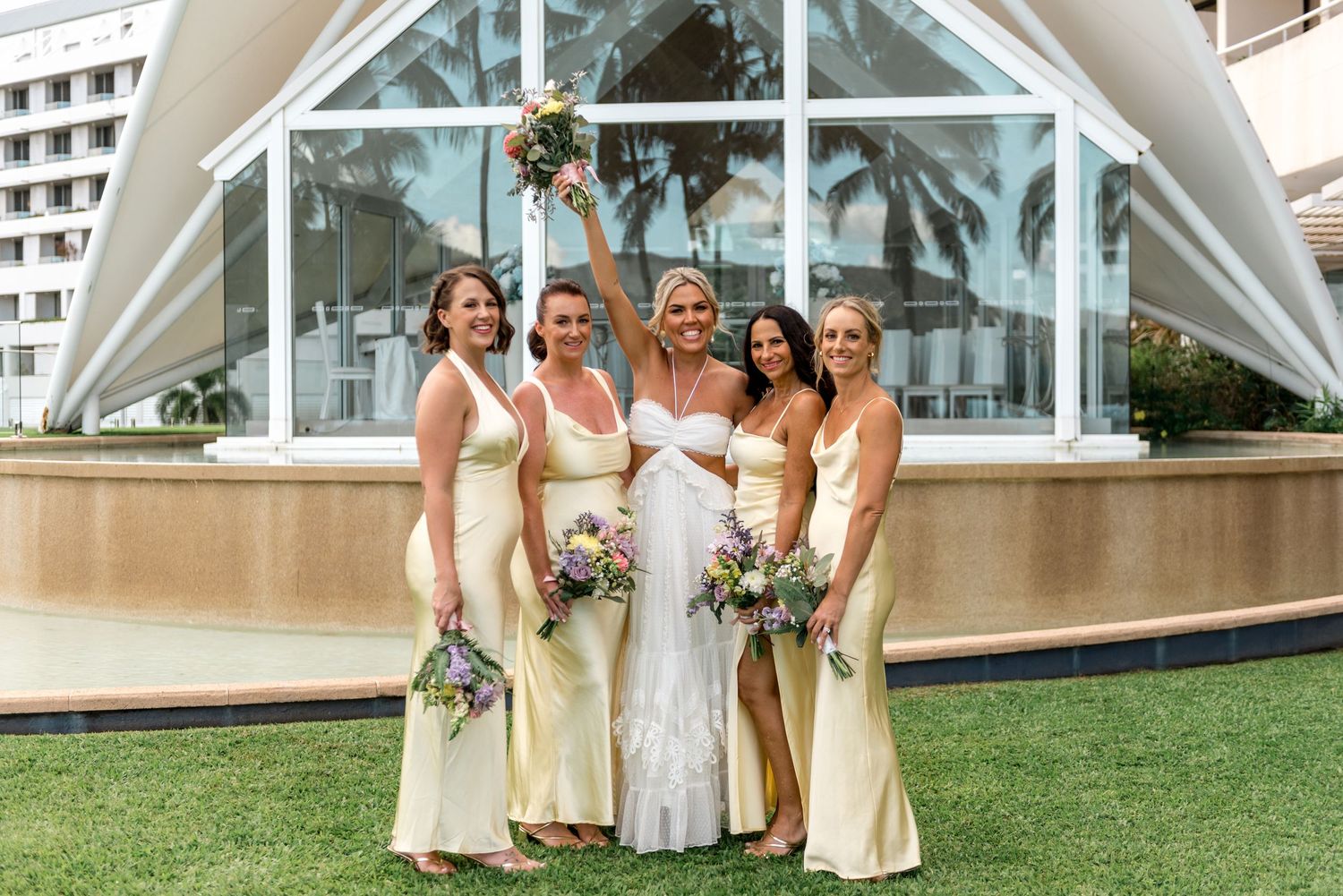 Bride in white gown celebrates with four bridesmaids in yellow dresses outside a glass chapel.