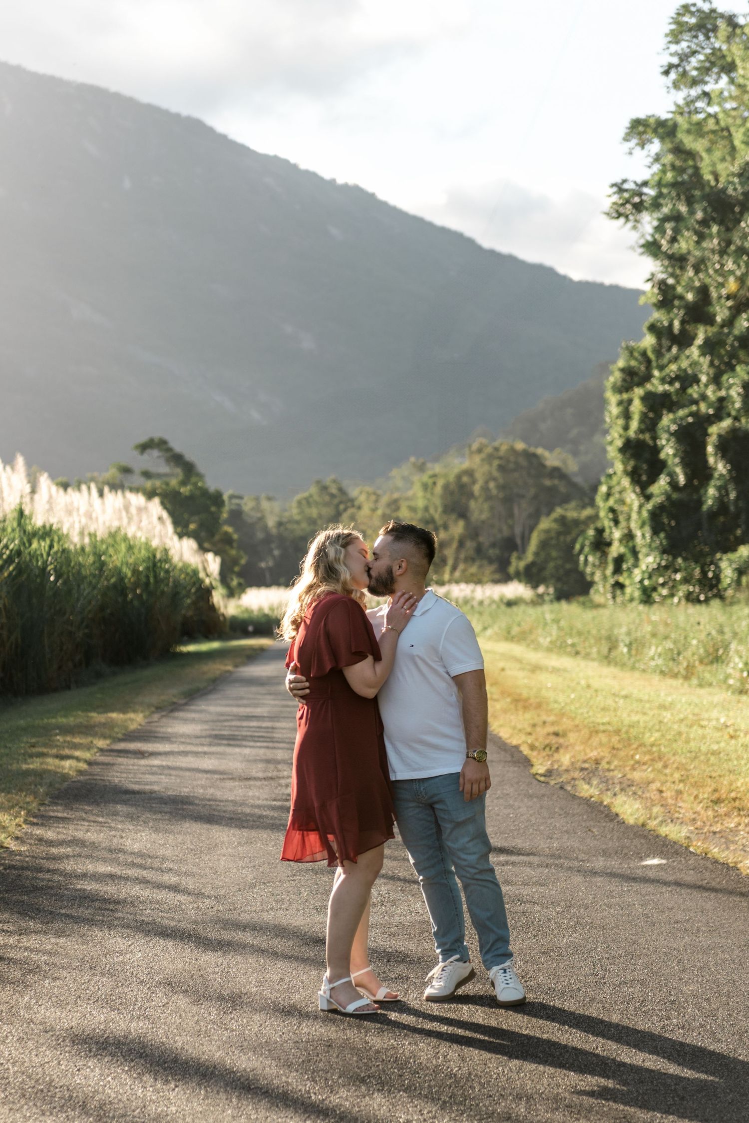 Romantic couple sharing a kiss on a sunlit rural road with lush green mountains in the background.