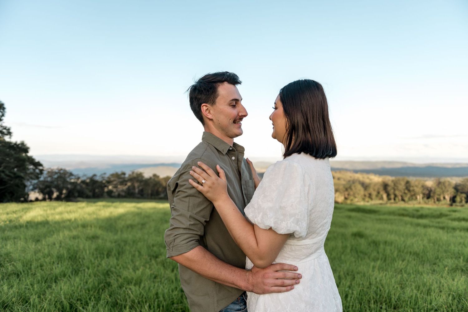 Couple embracing and smiling at each other in a lush green field at golden hour.