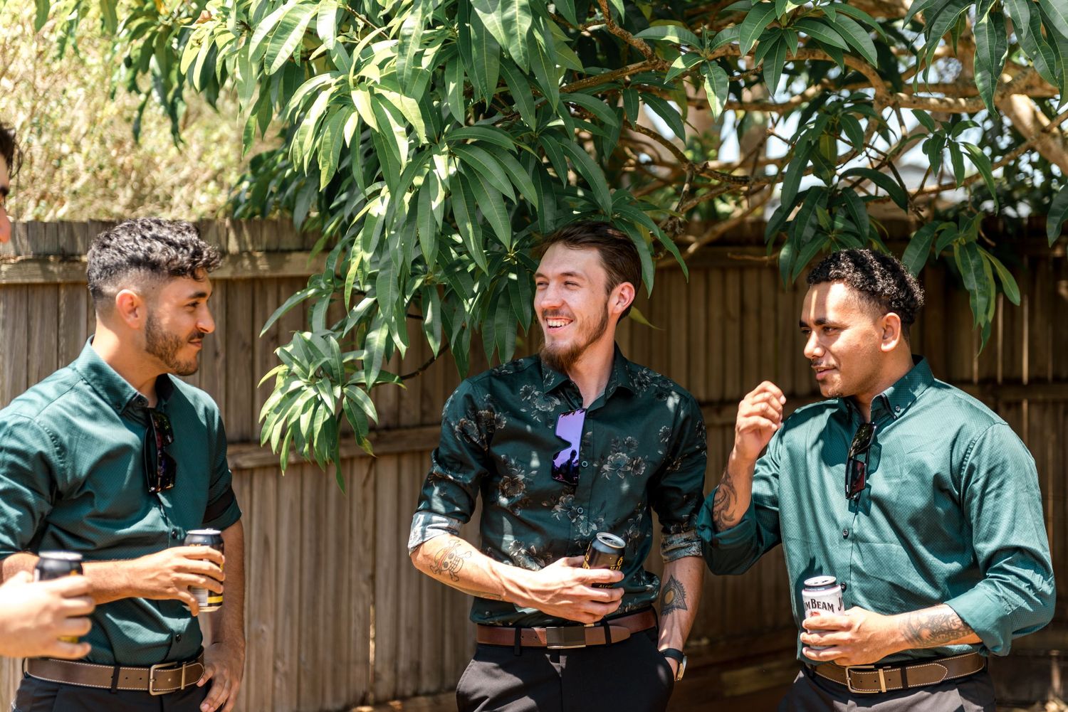 Three groomsmen in green shirts laughing and drinking coffee outdoors.